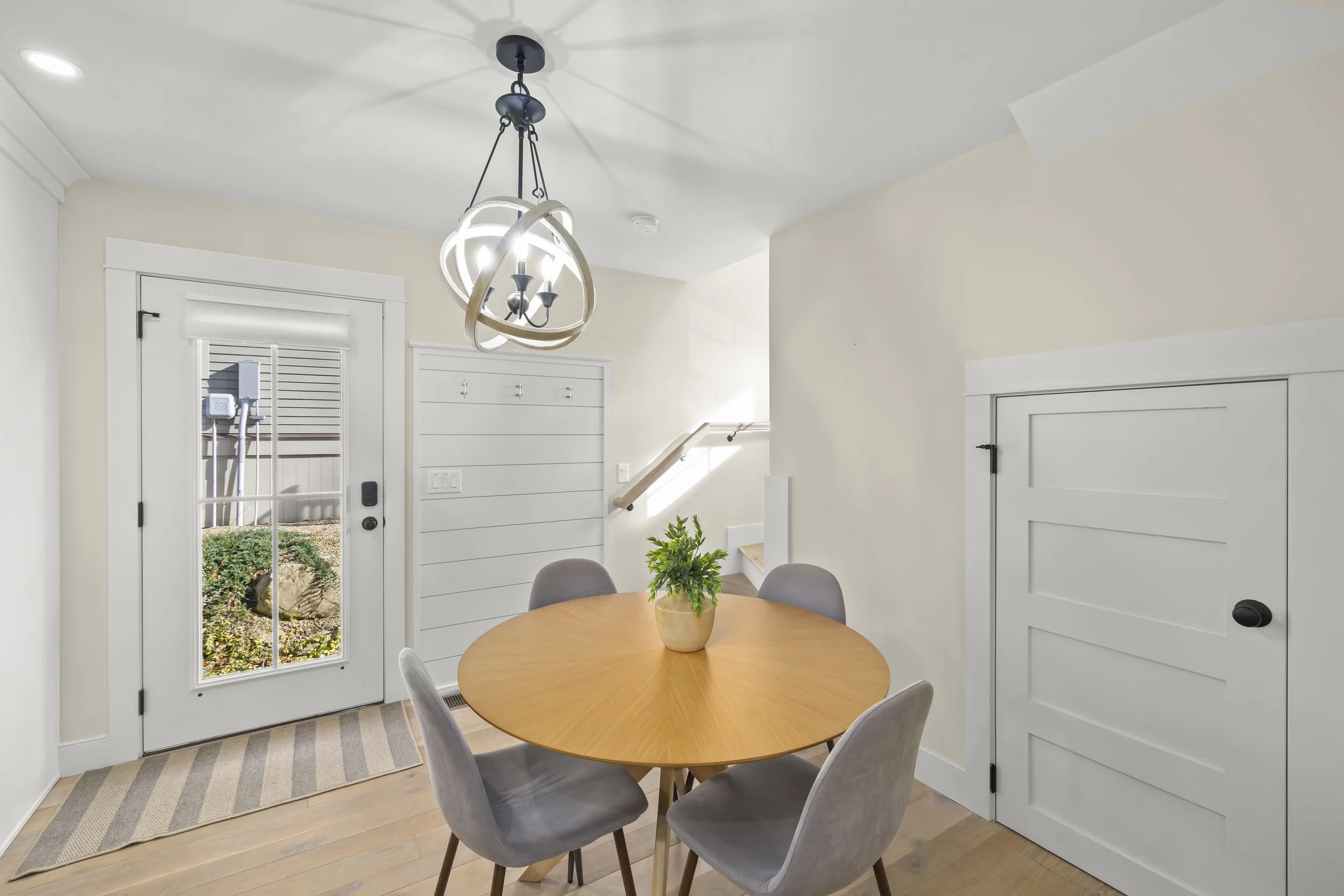 Dining area with a round wooden table, four gray chairs, and a plant centerpiece. A door with glass panels leads outside, and there is a small storage closet or cabinet with a white door. The walls are painted light, and a modern chandelier hangs fro