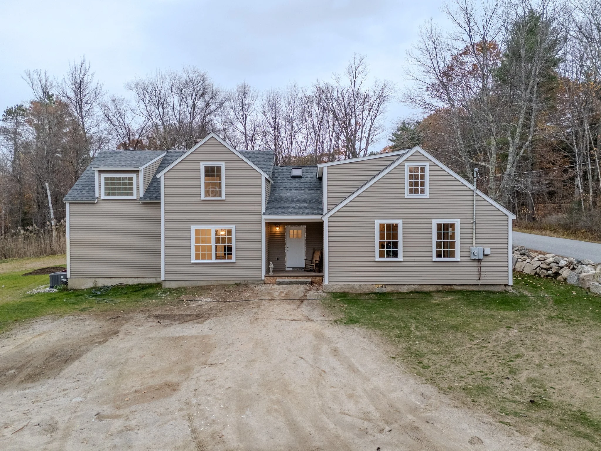 A two-story house with beige siding and white trim, featuring multiple windows and a covered porch in the front. The house is surrounded by trees with no leaves and is situated on a partly cleared lot with a dirt driveway and green grass.
