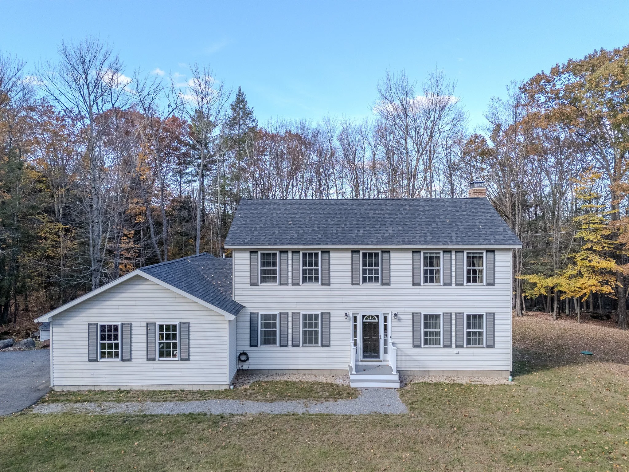 Front view of a two-story white house with black shutters, a gray roof, and a small set of stairs leading to the front door, surrounded by trees with some autumn foliage.