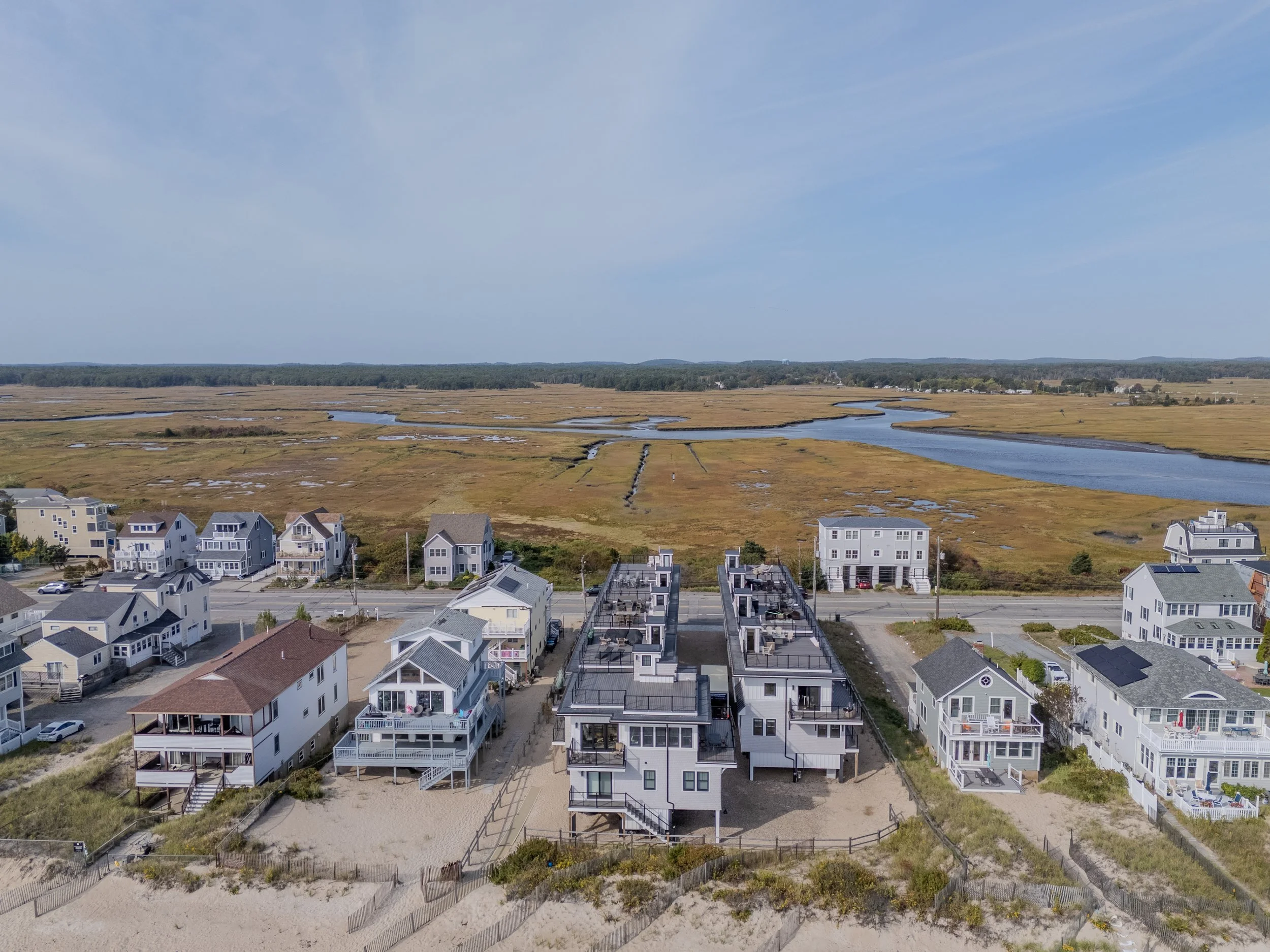 Aerial view of beach houses along the coast with marshland and water in the background under a partly cloudy sky.
