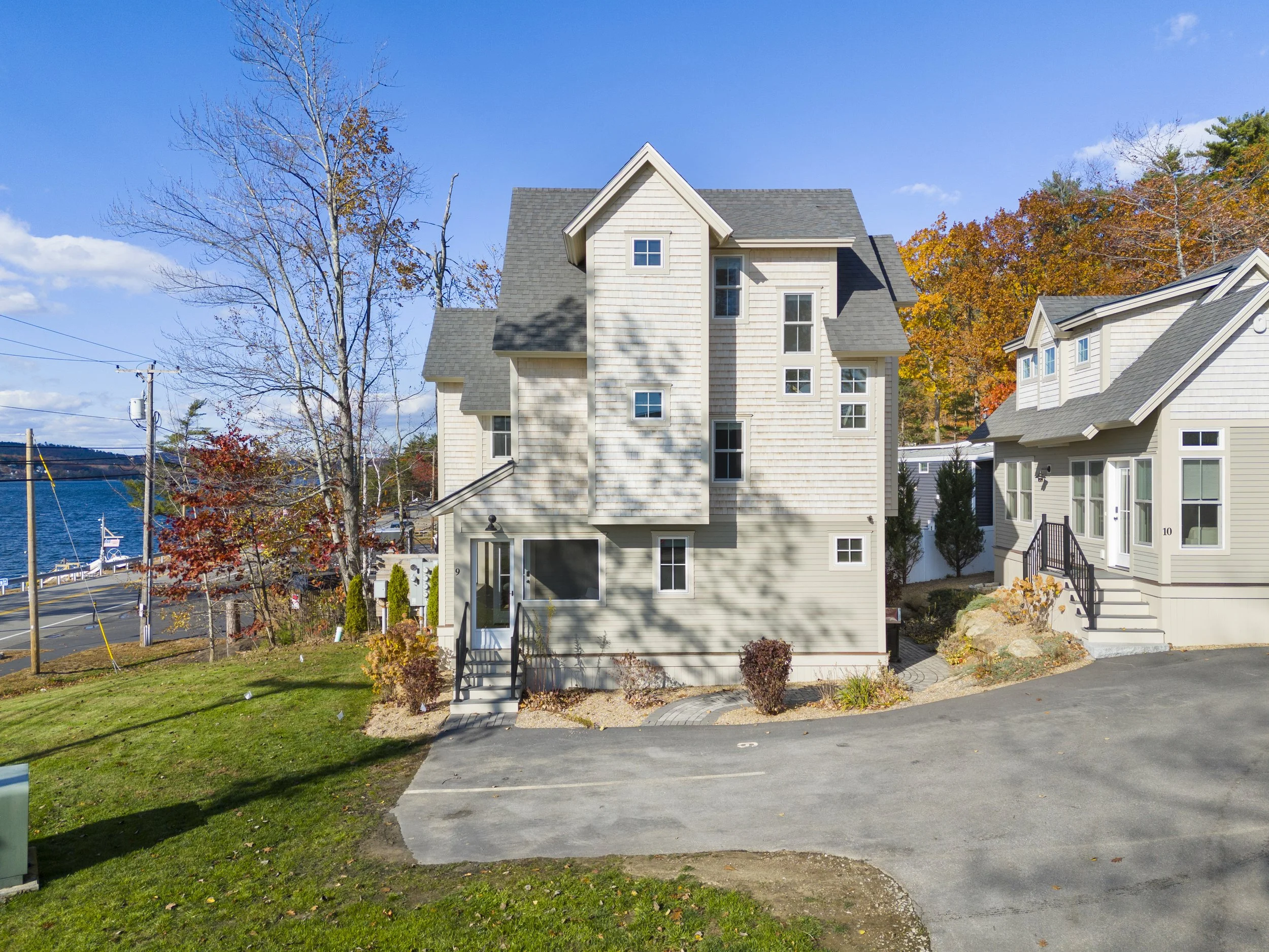 A modern house with multiple levels, light-colored siding and a dark roof, surrounded by a yard with green grass and bushes, near a body of water with trees displaying fall foliage in the background.