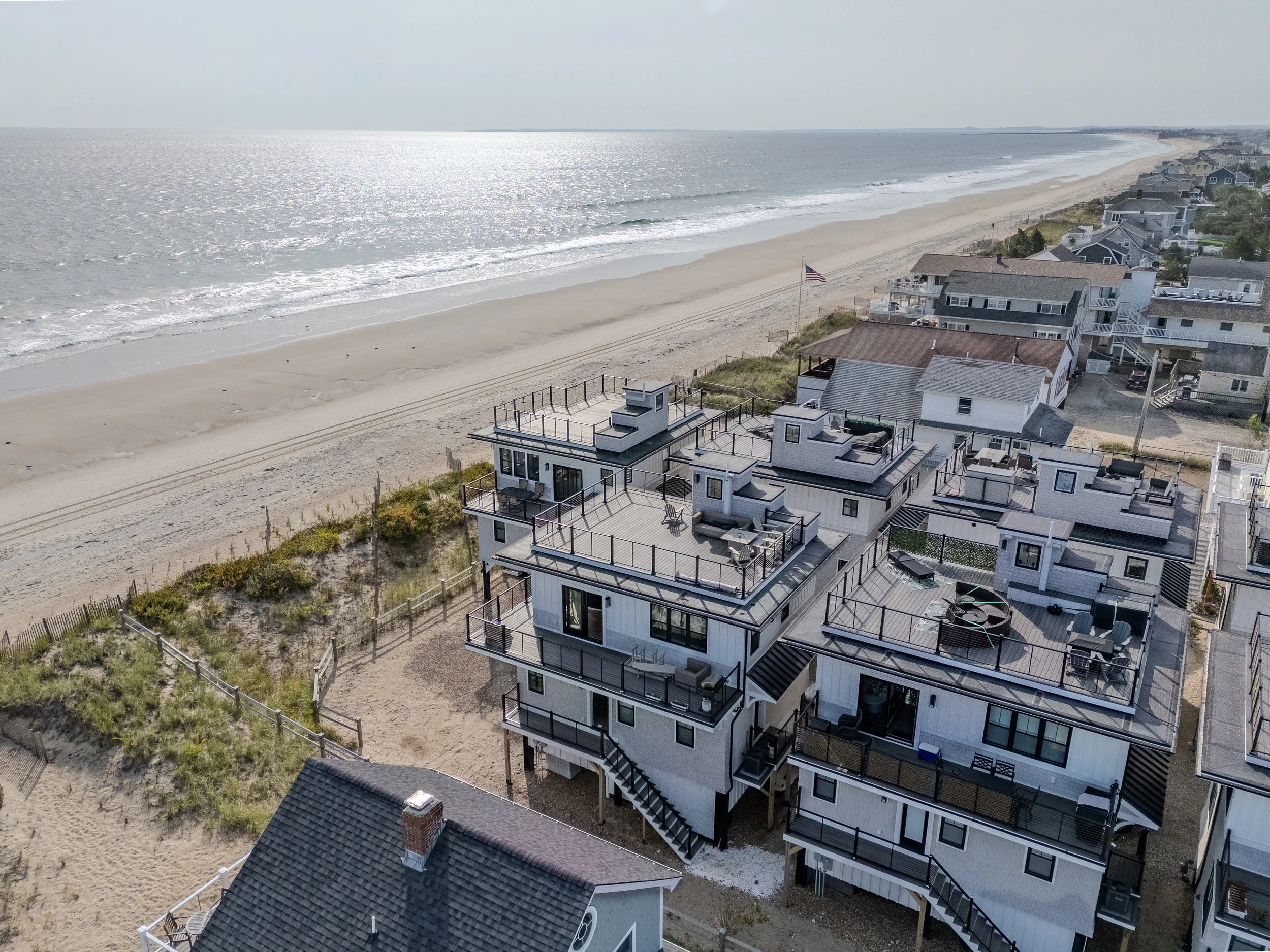 Aerial view of coastal houses along a sandy beach with ocean waves, some houses with rooftop decks and outdoor furniture, and an American flag flying.