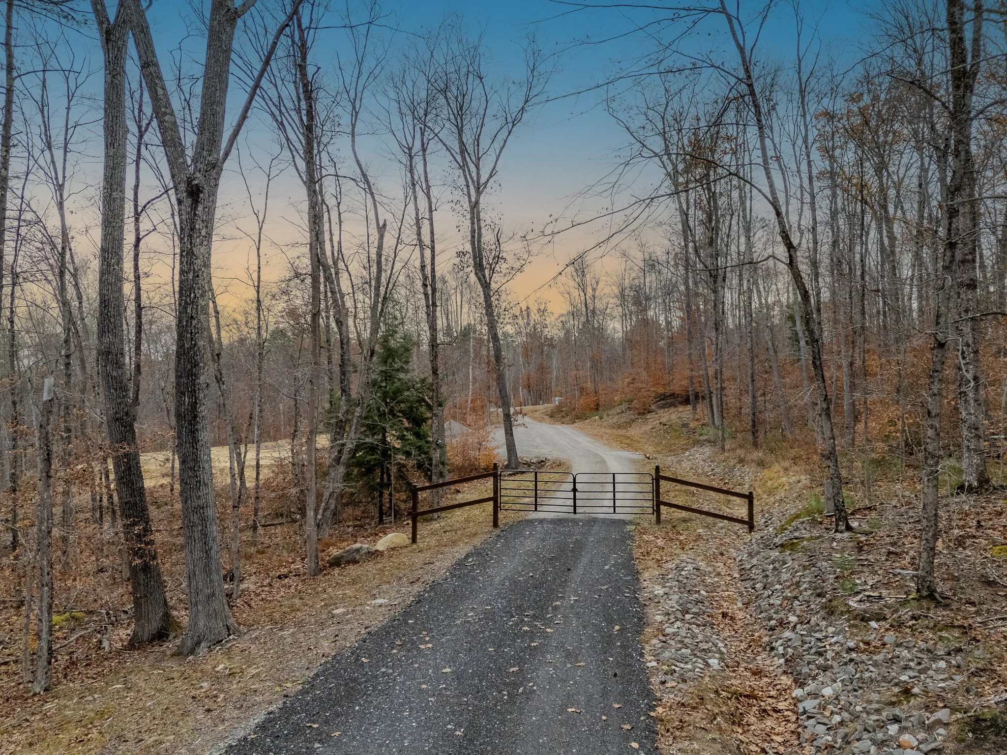 A gravel driveway leading through a wooded area with bare trees, a small wooden gate, and a dirt road stretching into the distance at sunset.
