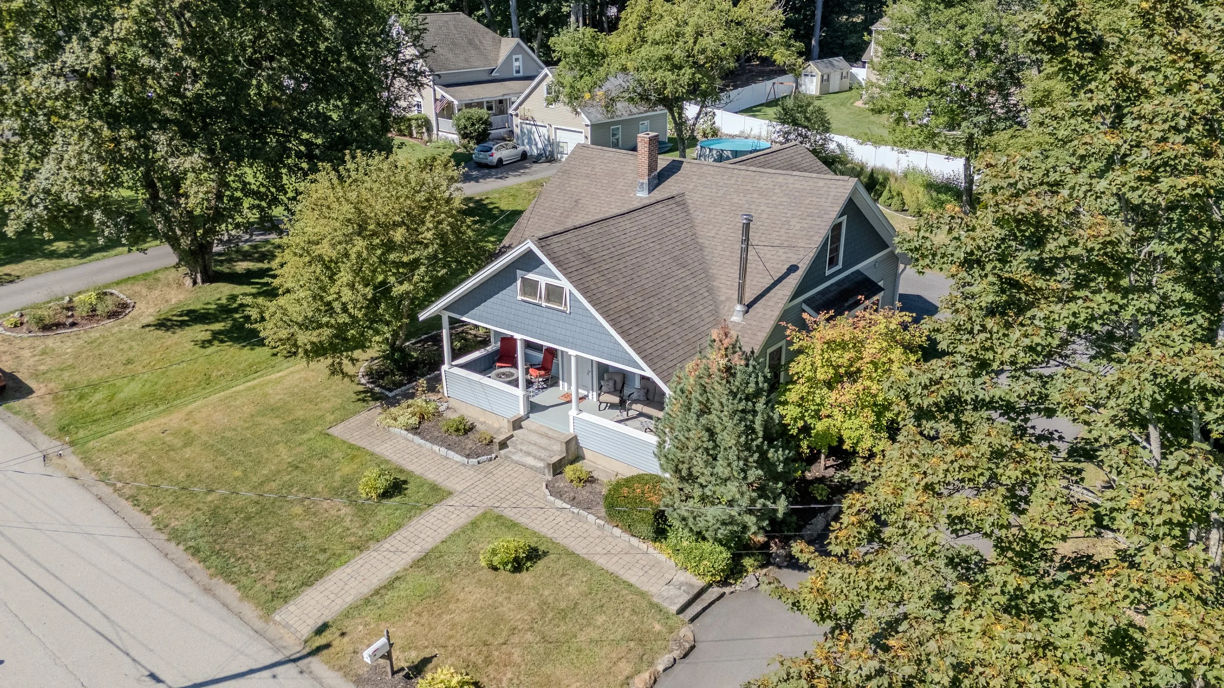 Aerial view of a blue house with a porch, surrounded by trees and a lawn, in a suburban neighborhood.
