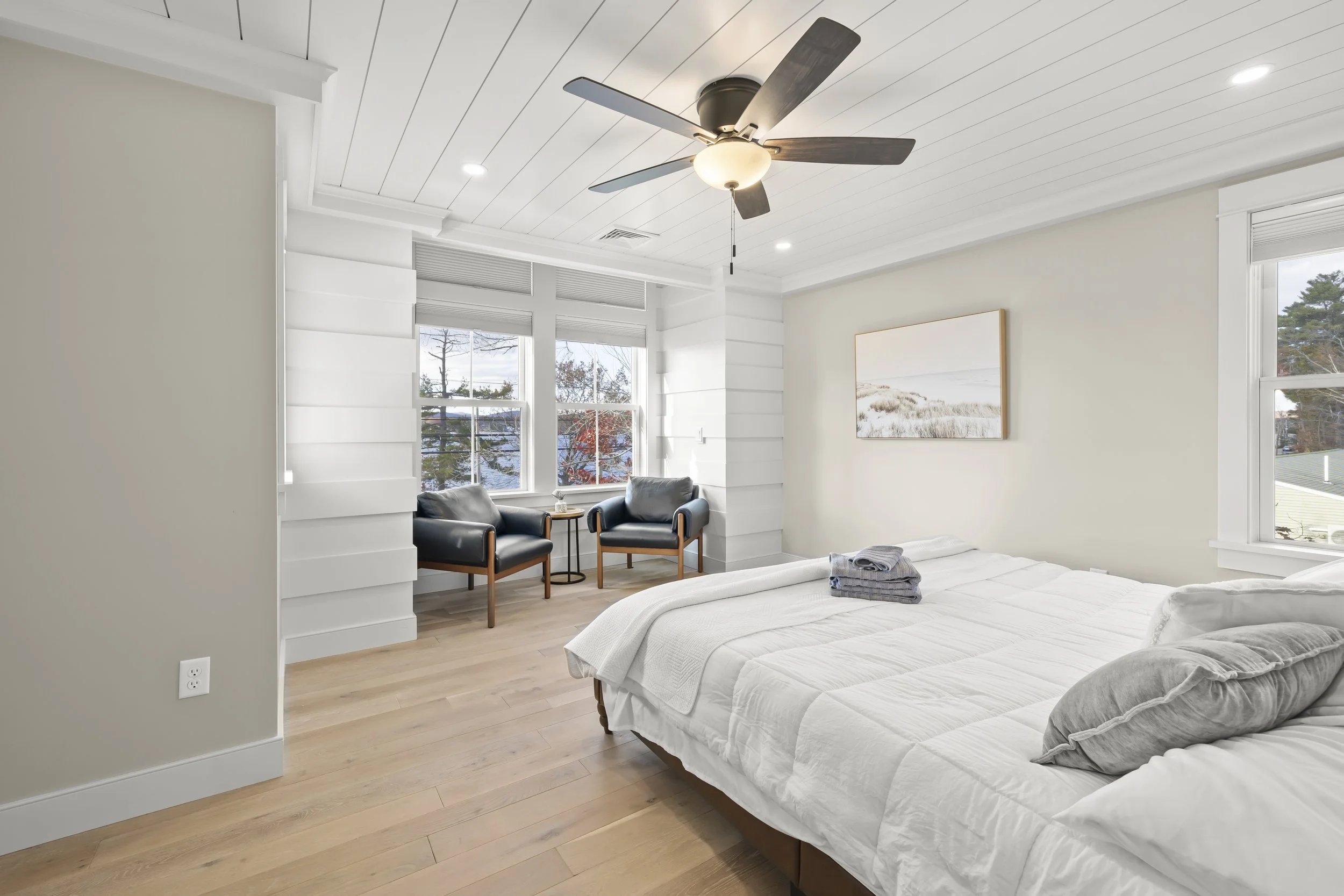 Bright bedroom with light wood floors, white walls, a ceiling fan, and two large windows. The bed has white bedding, and there are folded towels on it. Two black armchairs with wood accents sit by the windows, with a small side table between them. A 