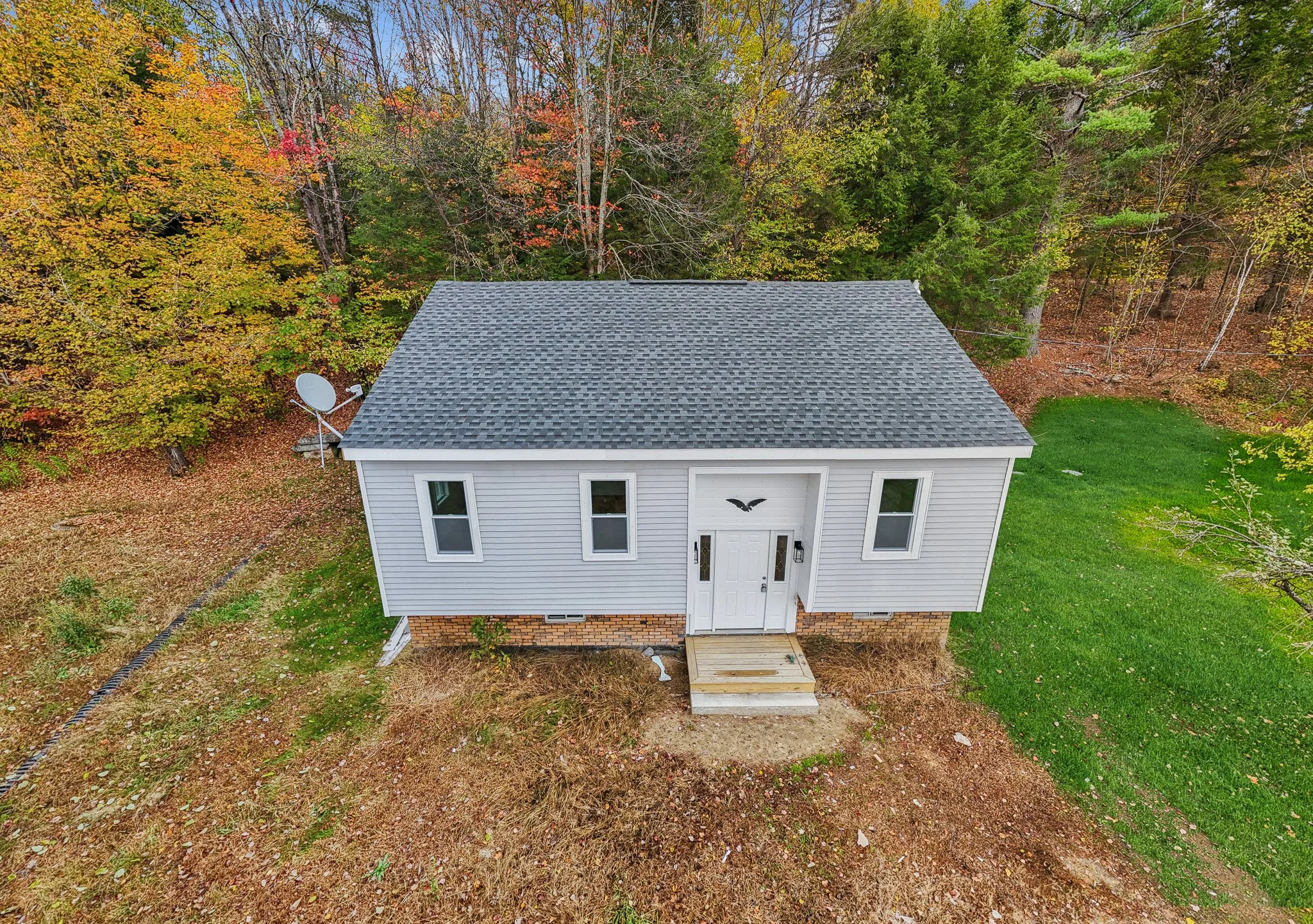 A small white house with a gray shingled roof, three windows on the front, a front door with a decorative bat silhouette above it, and a satellite dish on the left side. The house is surrounded by trees with autumn leaves and a green lawn on the righ
