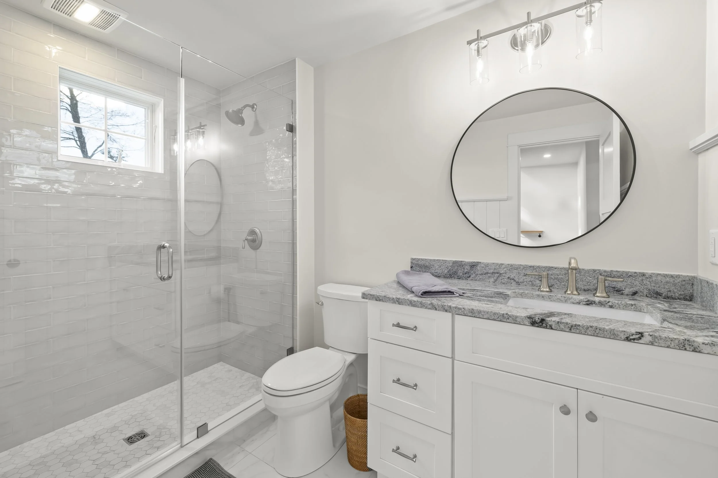 Modern bathroom with white tile shower, white toilet, gray granite vanity with sink, round mirror, and overhead lighting