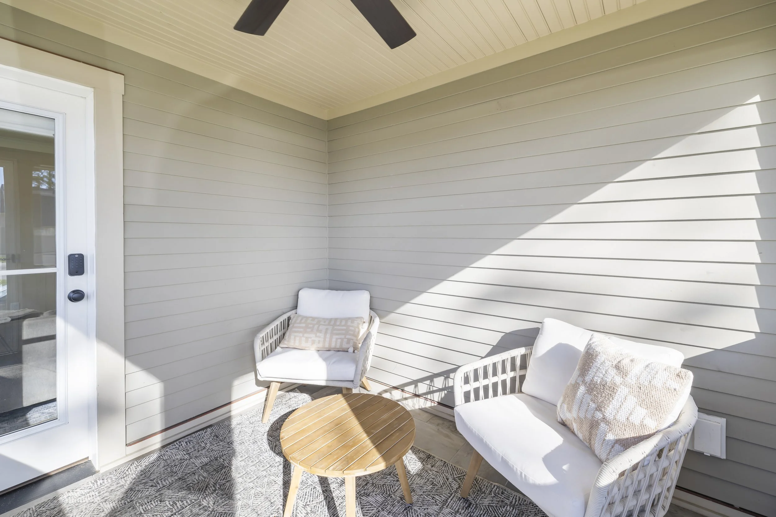 Patio area with two white cushioned chairs, a small round wooden table, and a gray patterned rug, with a sliding glass door and beige siding wall.