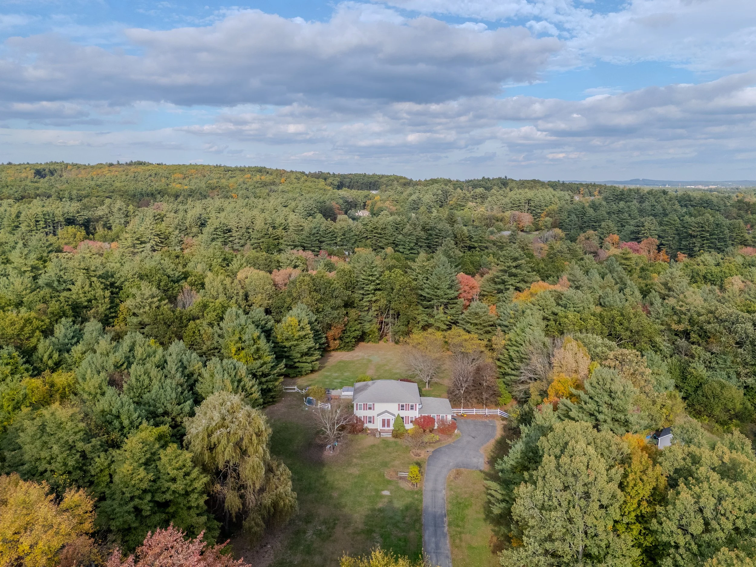 Aerial view of a white house with a gray roof, surrounded by trees with fall foliage and a curved driveway leading to it, in a forested landscape under a partly cloudy sky.