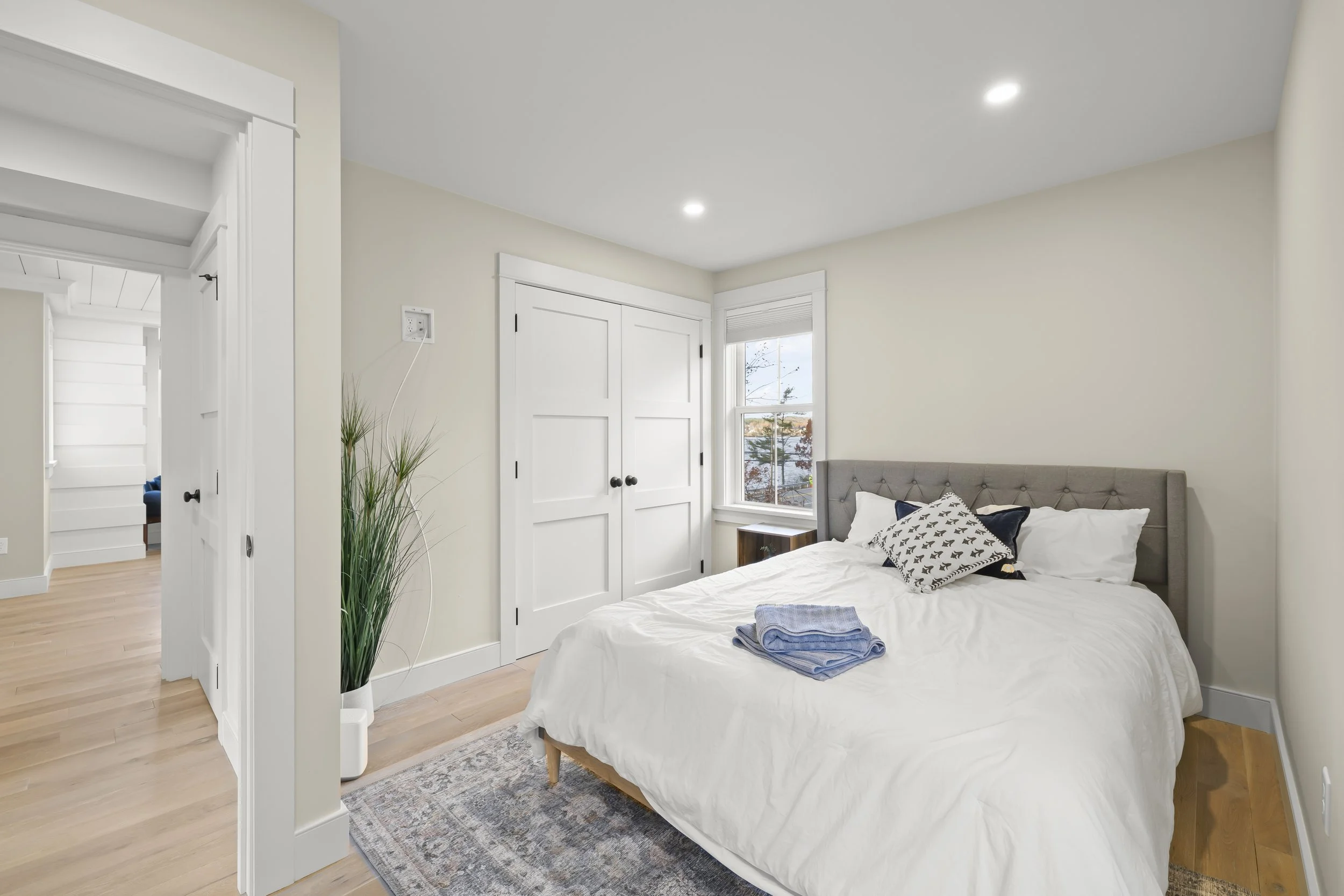 A clean, well-lit bedroom with white walls, a gray bed with pillows, a nightstand by the window, and a folded set of blue towels on the bed.