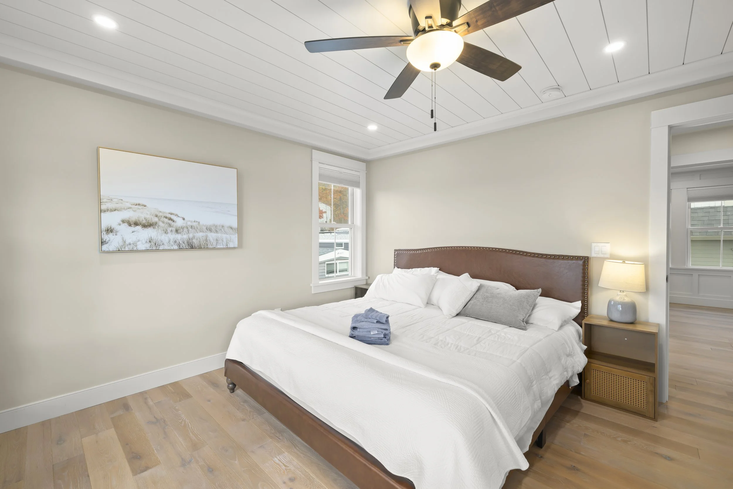 A bedroom with a large bed, white bedding, and pillows, next to a nightstand with a lamp, and a wall painting of a beach scene. There is a window revealing a neighboring house, and a ceiling fan with light.