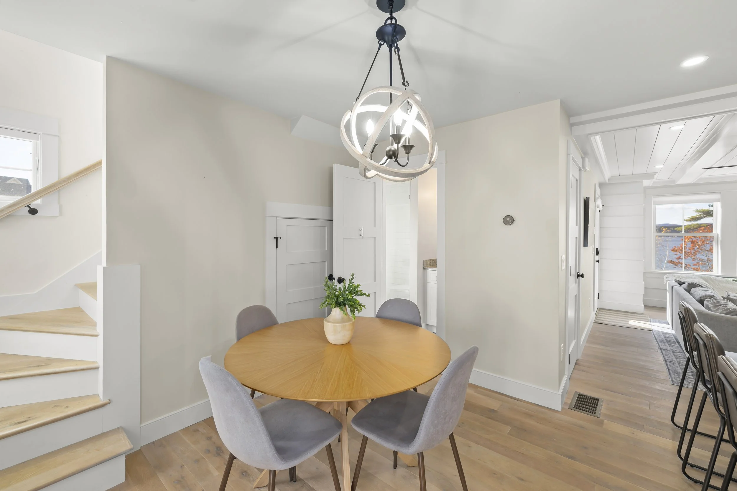 A dining area with a round wooden table, five gray chairs, and a modern chandelier. There is a potted plant in the center of the table. The background includes a staircase, a hallway, and a living room with large windows showing a view outside.