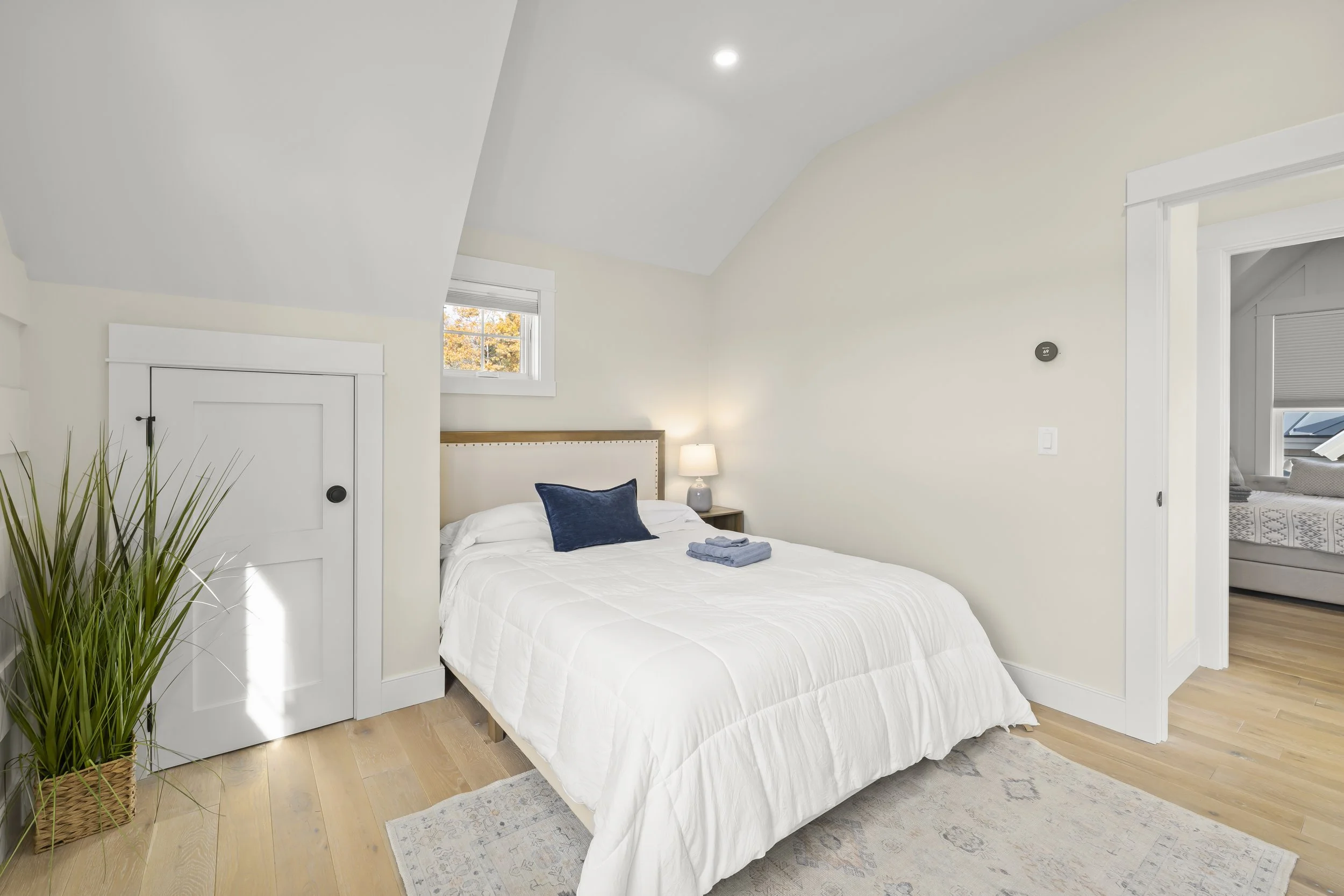 A cozy bedroom with a white bed, blue pillow, nightstand with lamp, small window, and a closet door, with hardwood floors and light-colored walls.