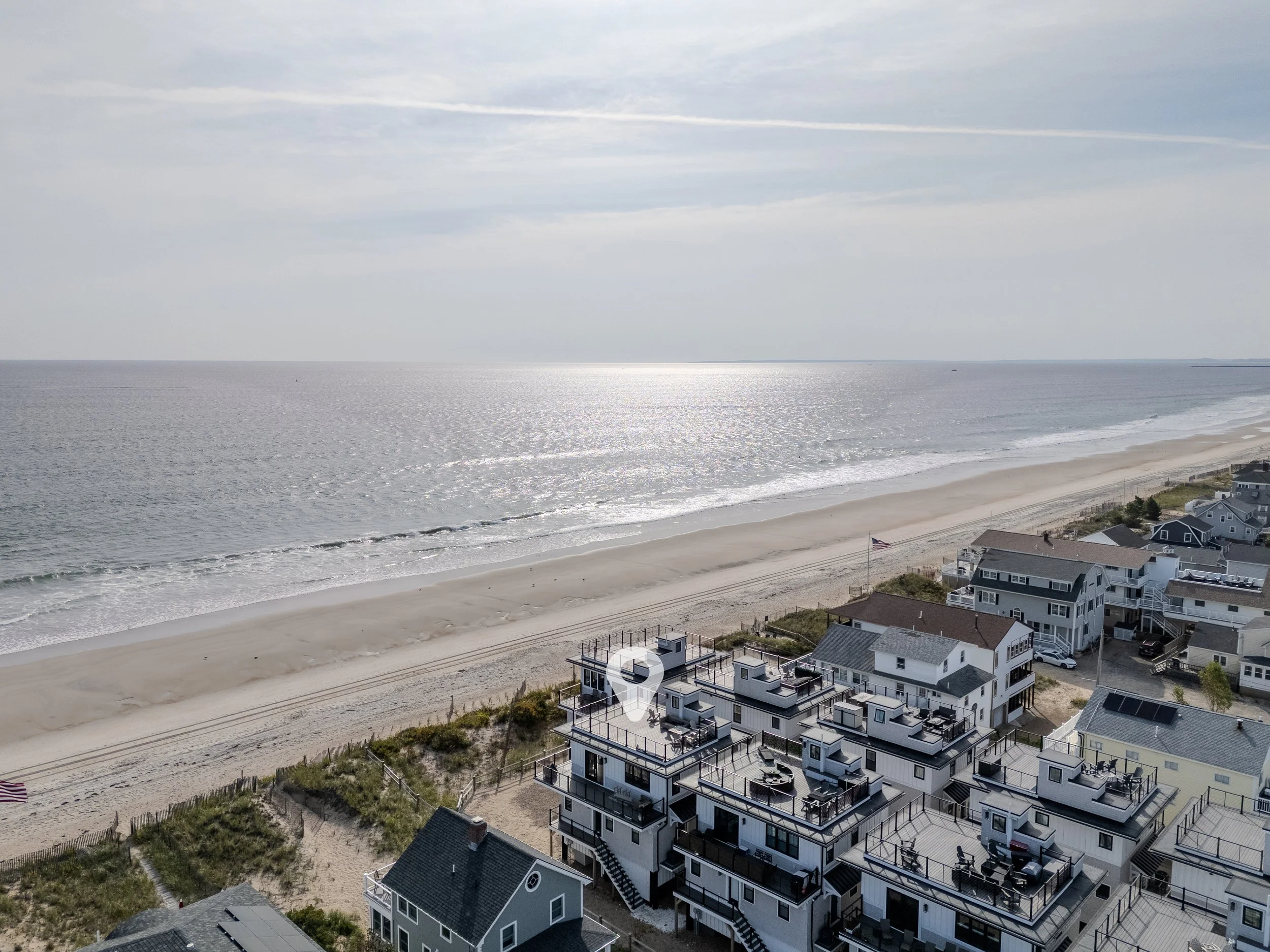 Bird's eye view of a beach with white sand, ocean waves, and a row of multi-story beach houses with decks facing the water. An American flag is flying near the houses.