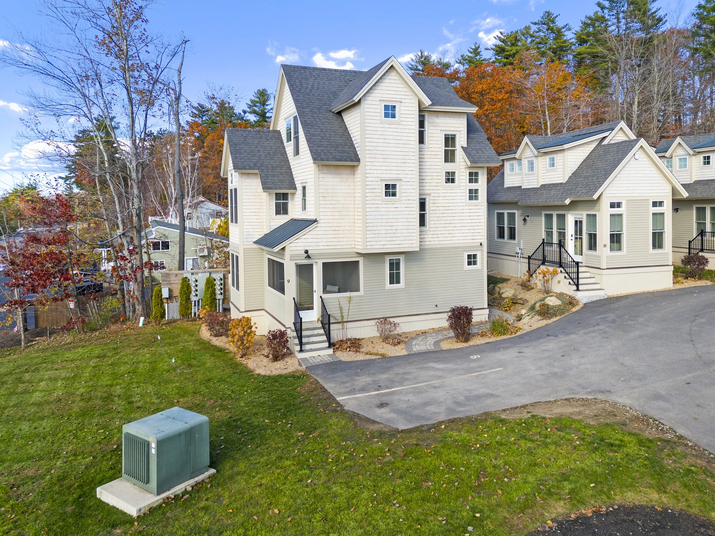 Multiple modern houses with beige siding, black railings, and stairs, set in a neighborhood with trees showing fall colors, on a partly cloudy day.