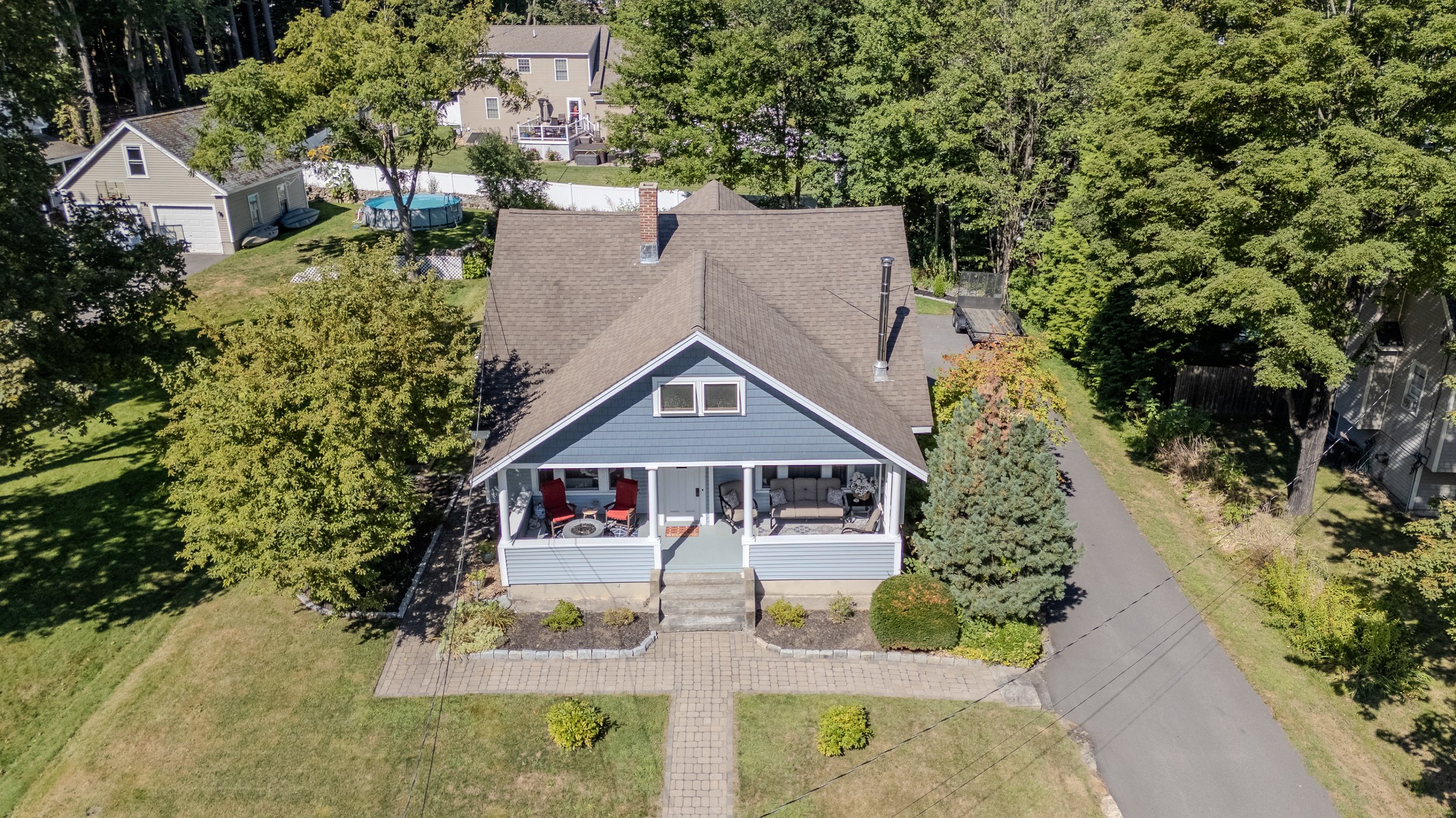 Aerial view of a blue house with a covered front porch, surrounded by trees and a lawn, with neighboring houses and a driveway visible.
