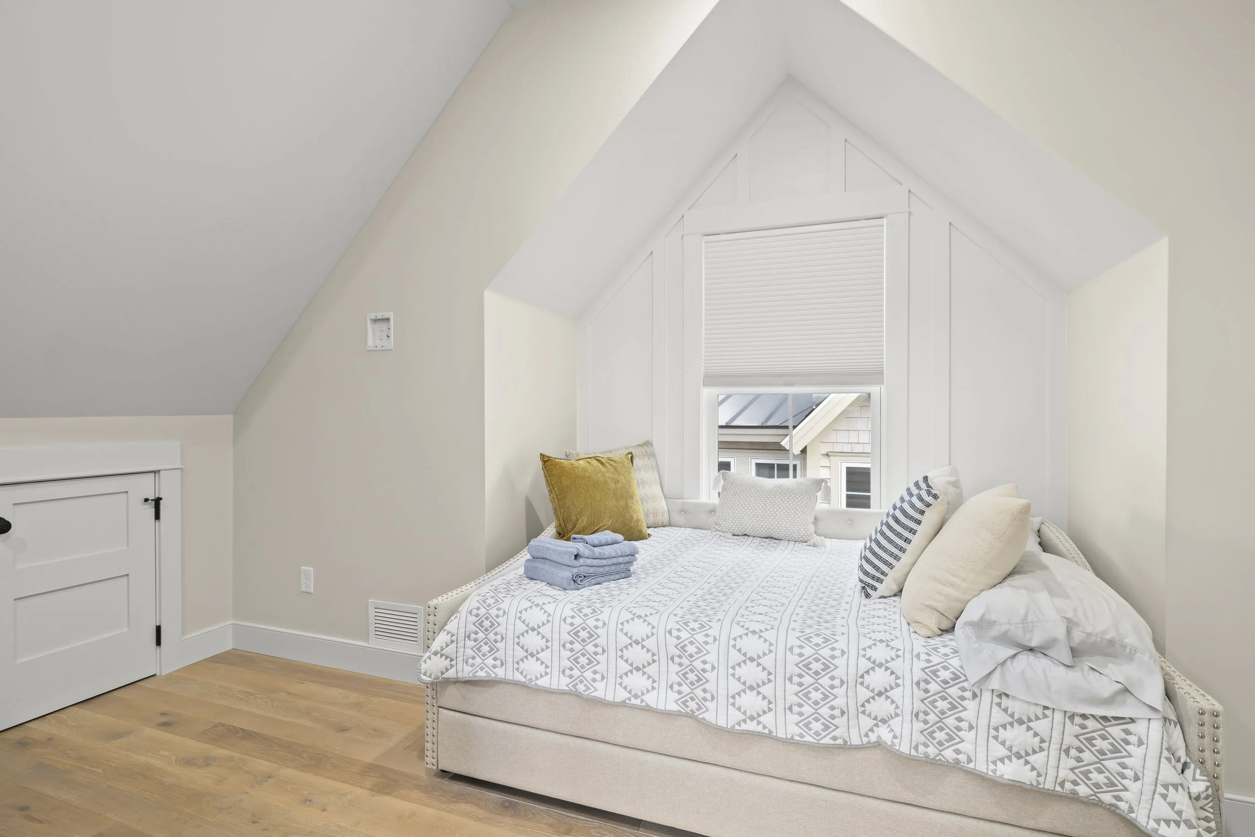 A cozy bedroom with white walls and sloped ceiling, featuring a bed with white and beige bedding, pillows, and a small window with closed blinds, overlooking neighboring houses.