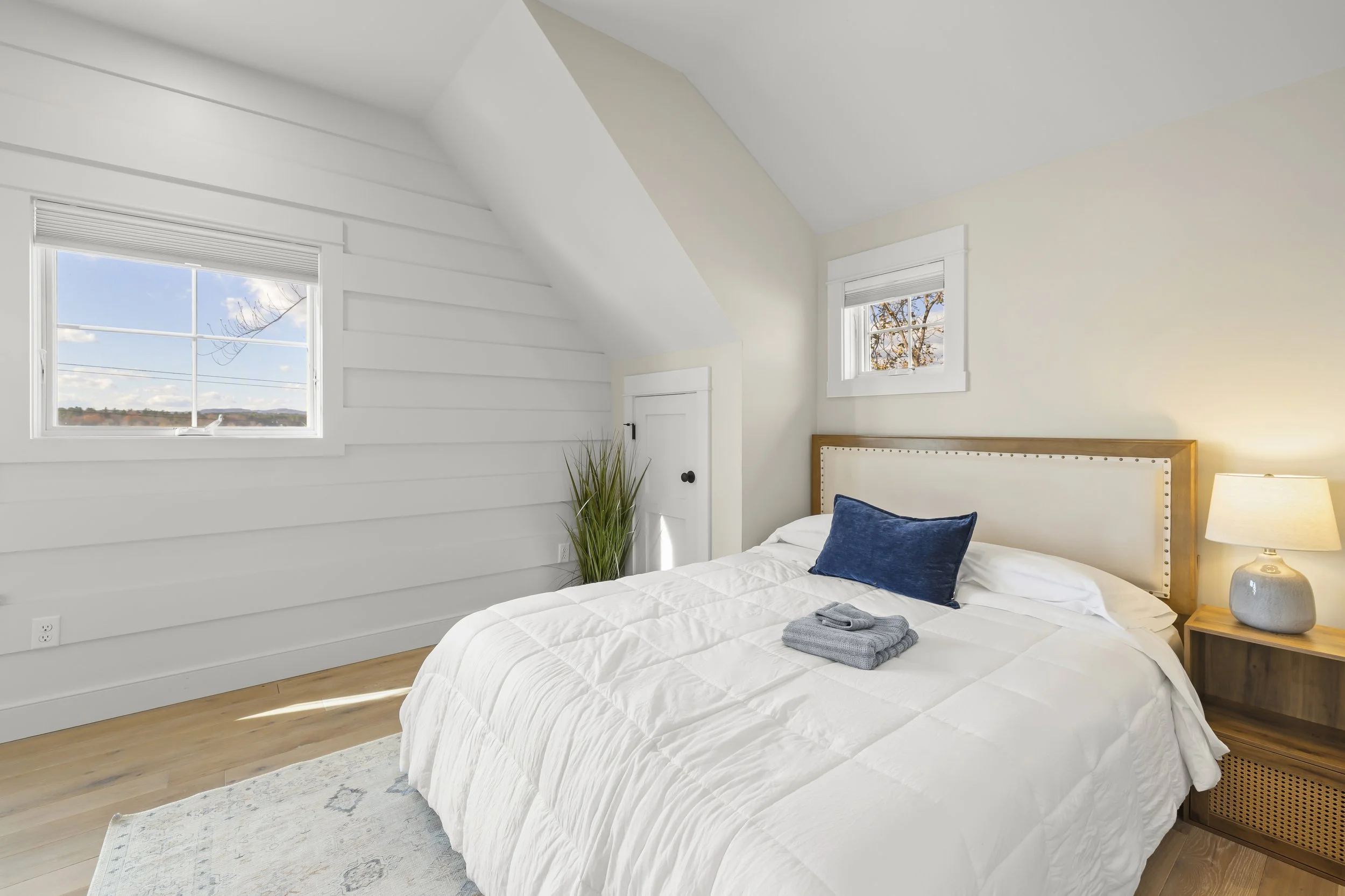 A bright bedroom with white walls, light hardwood floors, a bed with white bedding and a blue pillow, a bedside table with a lamp, and two small windows showing a view of trees and sky.