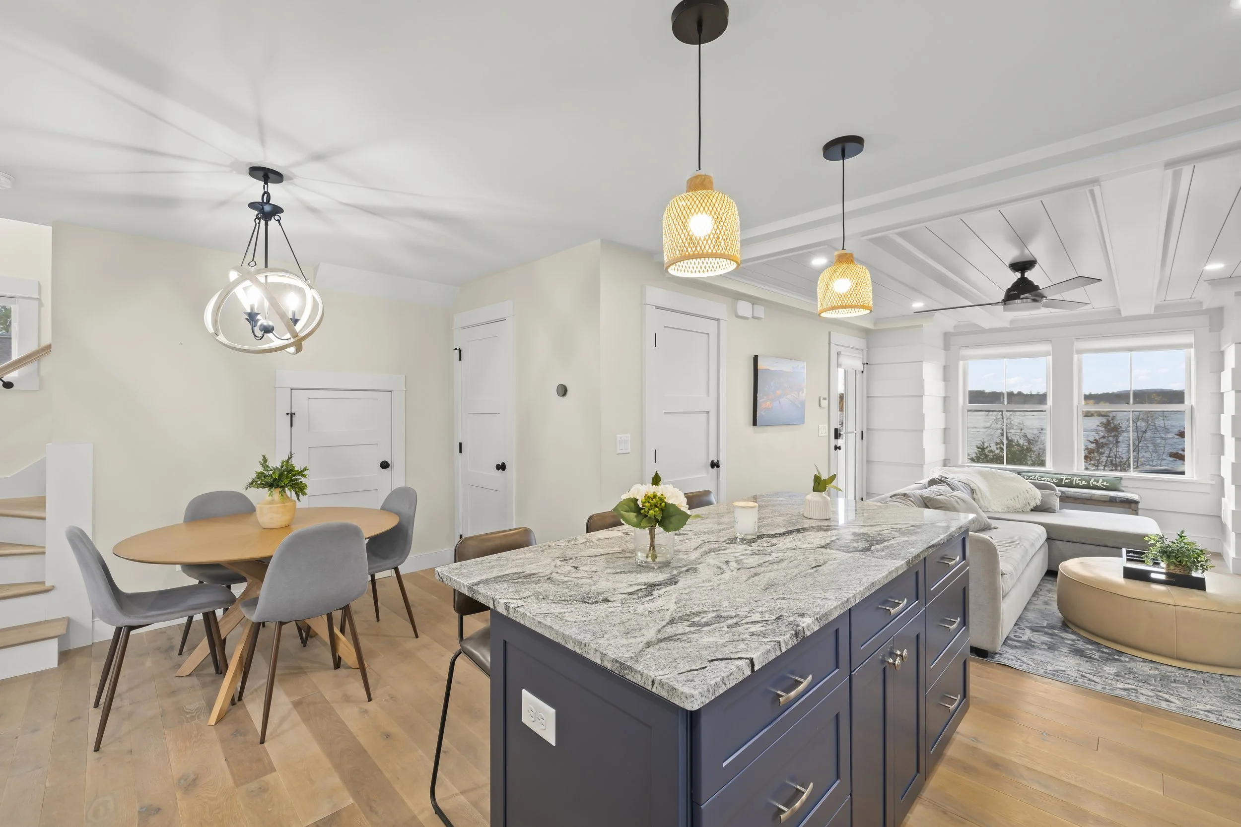 Open-concept kitchen and living room with a granite island, a dining table with four chairs, a sofa, a wall-mounted TV, large windows overlooking a lake, and pendant and ceiling lights.