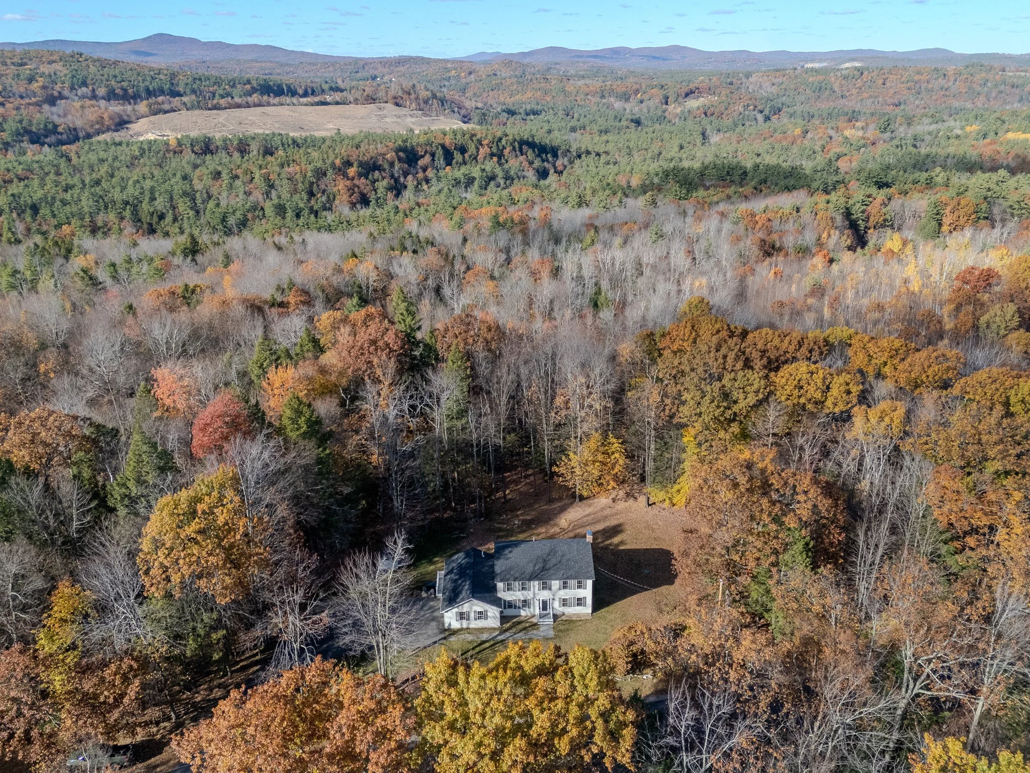 Aerial view of a house surrounded by a forest in autumn with trees showing fall colors, and distant mountains in the background.