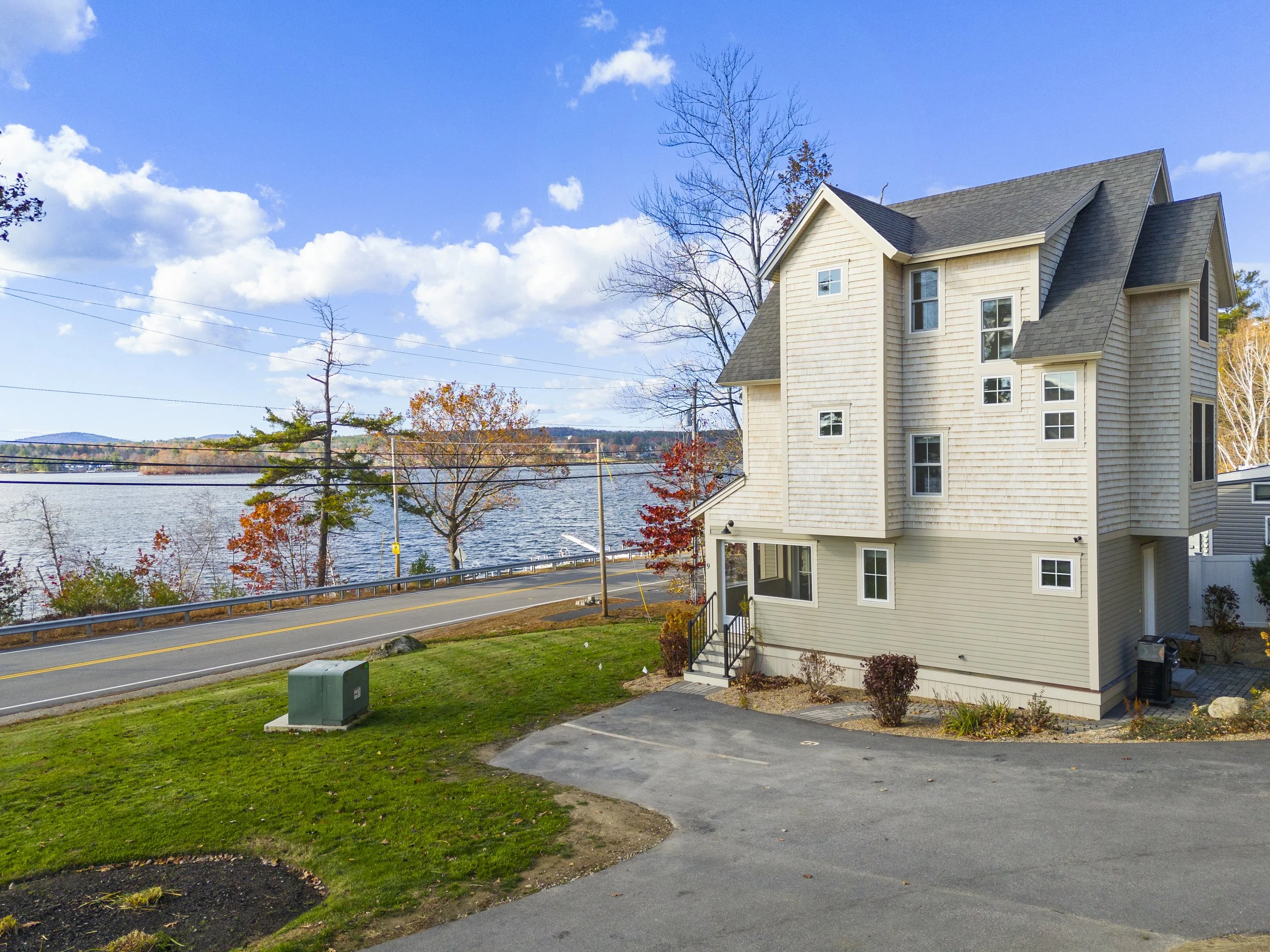 A beige multi-story house situated near a body of water with trees showing fall foliage and a clear blue sky.