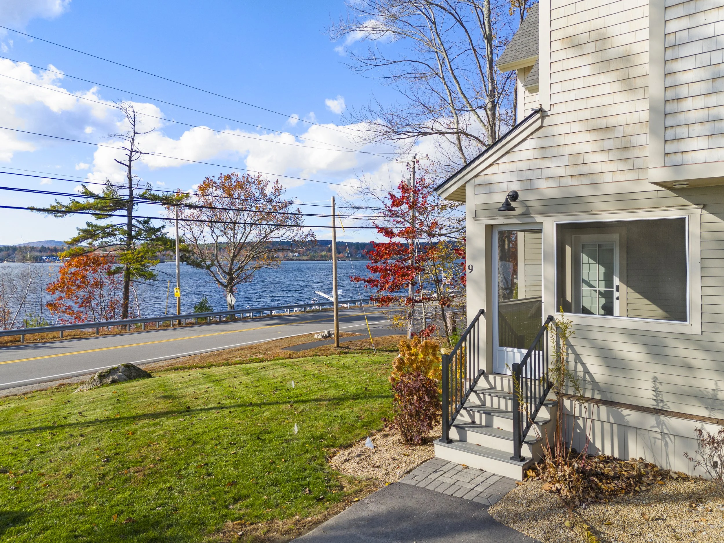 View of a house with a small porch and steps near a waterfront, with trees displaying autumn colors, a road, and a body of water in the background under a partly cloudy sky.
