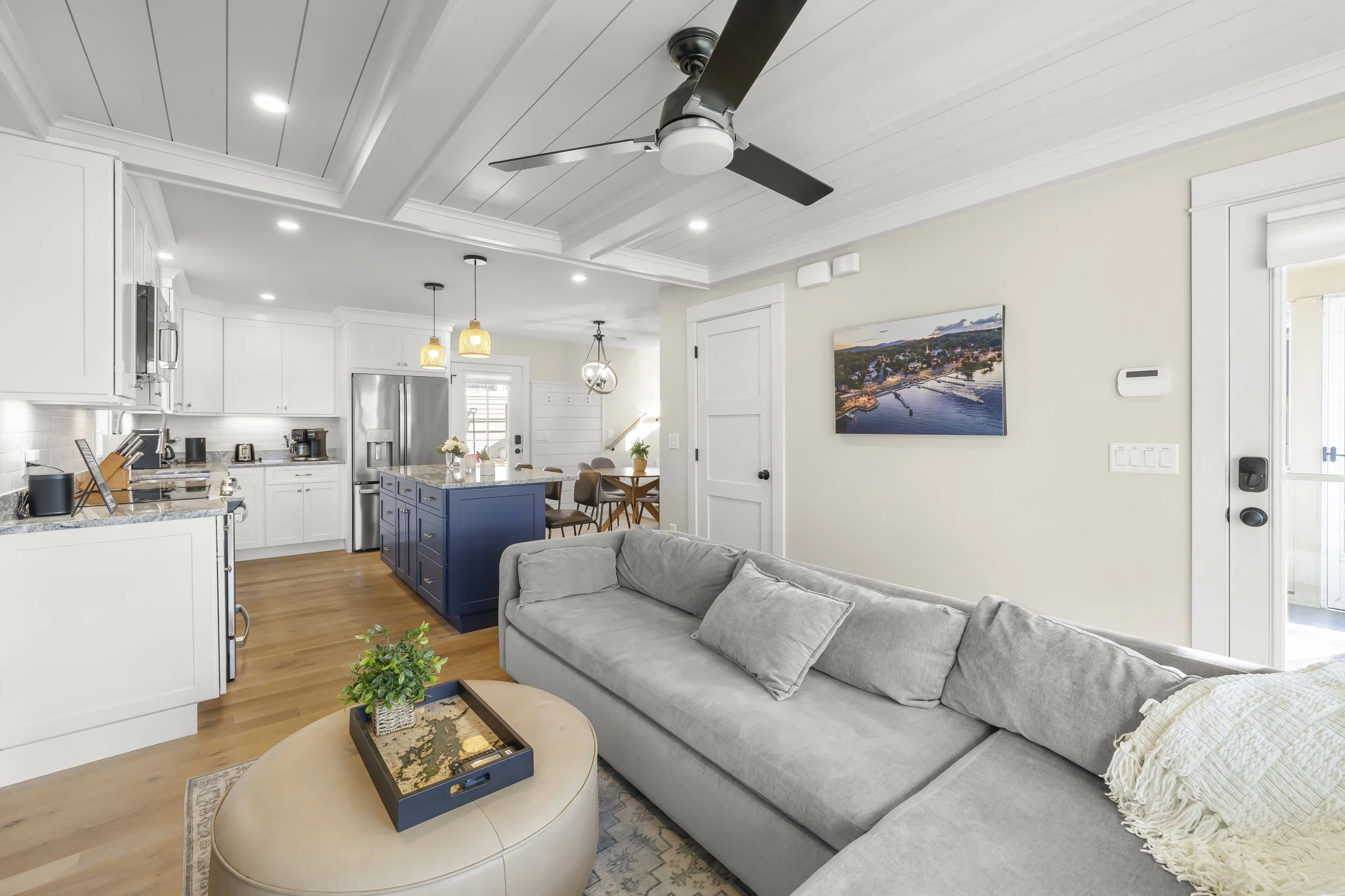 Open concept living room and kitchen in a modern home. The living area has a light gray sofa, a coffee table with a small plant, and a wall-mounted television. The kitchen features white cabinets, a blue island, stainless steel refrigerator, and pendant lighting. The space has hardwood flooring and a white beamed ceiling with a ceiling fan.