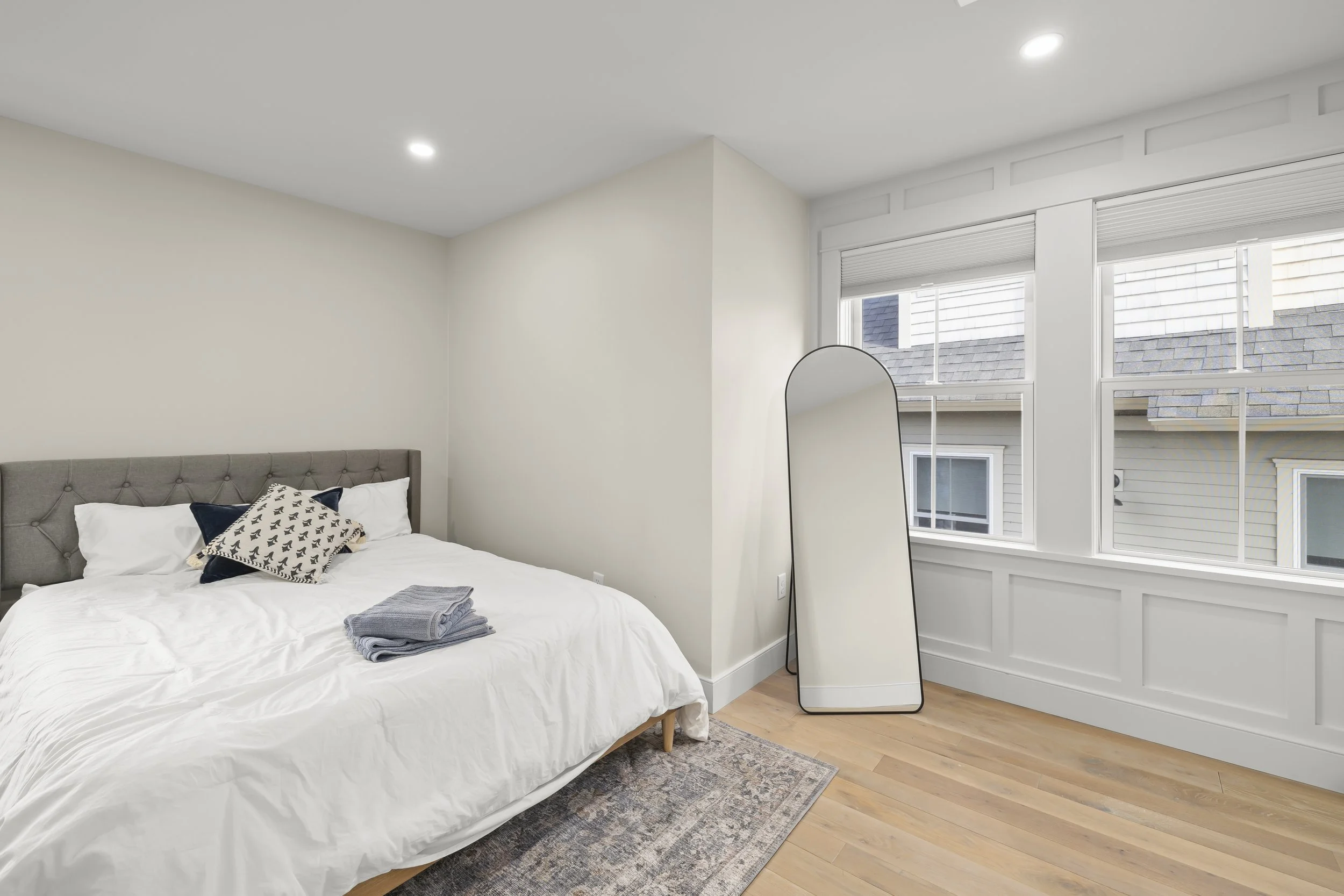 A bedroom with a bed, a mirror, and a large window, featuring hardwood flooring and a neutral color scheme.