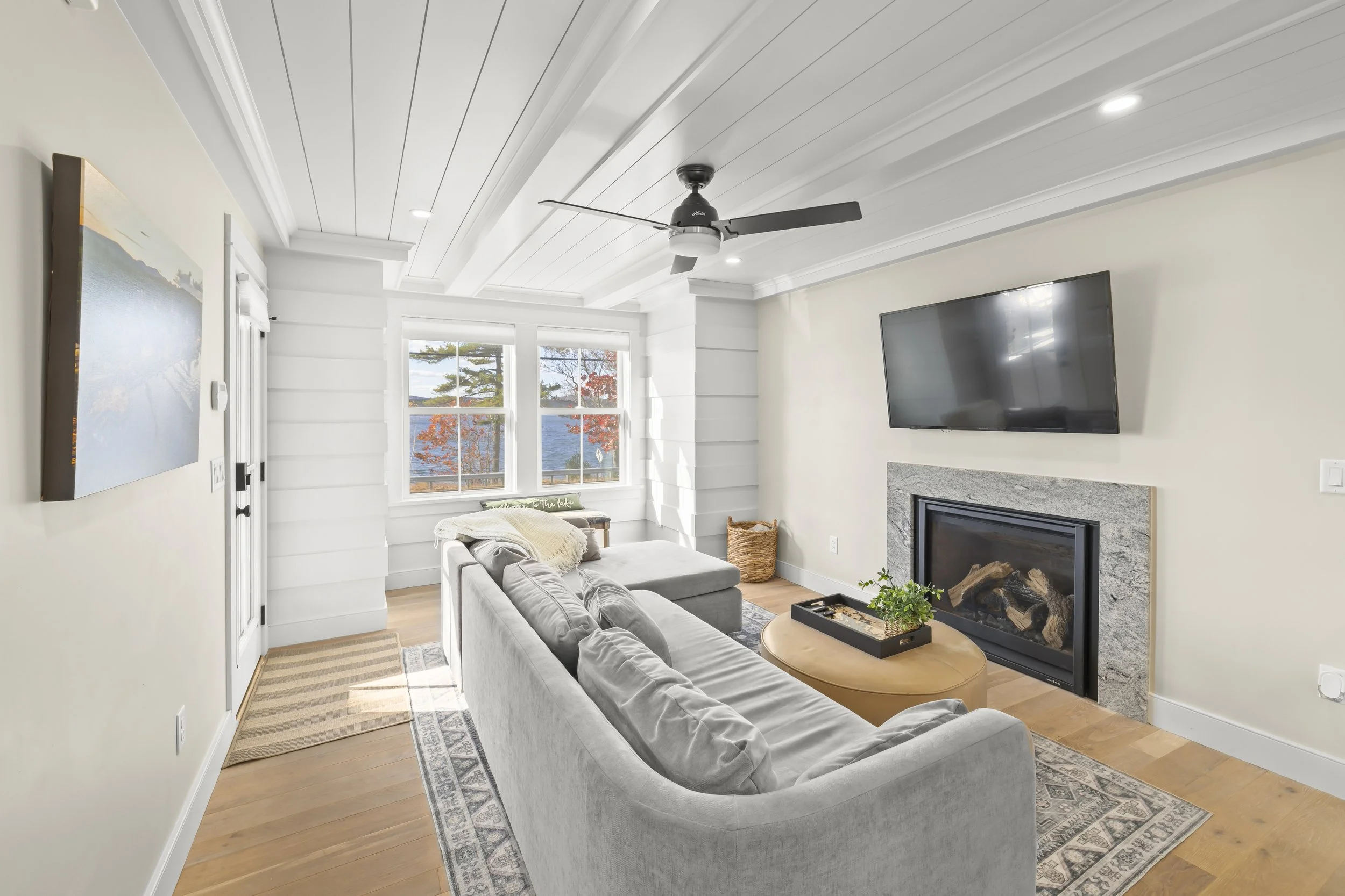 Living room with white walls, ceiling, and trim, gray sofa with pillows, oval coffee table with plant, fireplace with granite surround, mounted TV, large window showing trees and lake, ceiling fan, hardwood floors, and a rug.