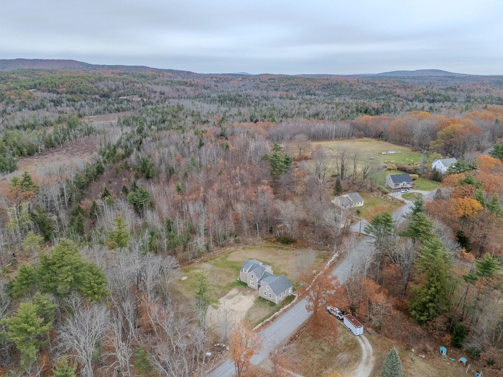 Aerial view of a rural landscape with scattered houses, a dirt driveway, and dense trees with fall foliage in the background.