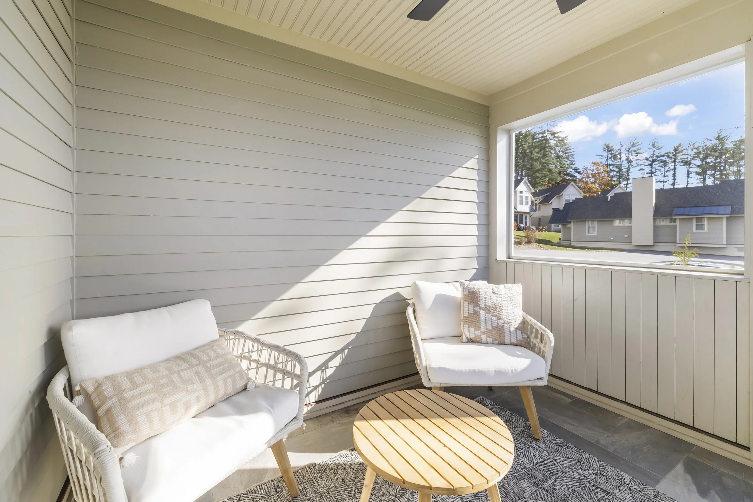 Sunlit porch with white wicker chairs, cushions, a small round wooden table, and a large window showing neighboring houses and trees.