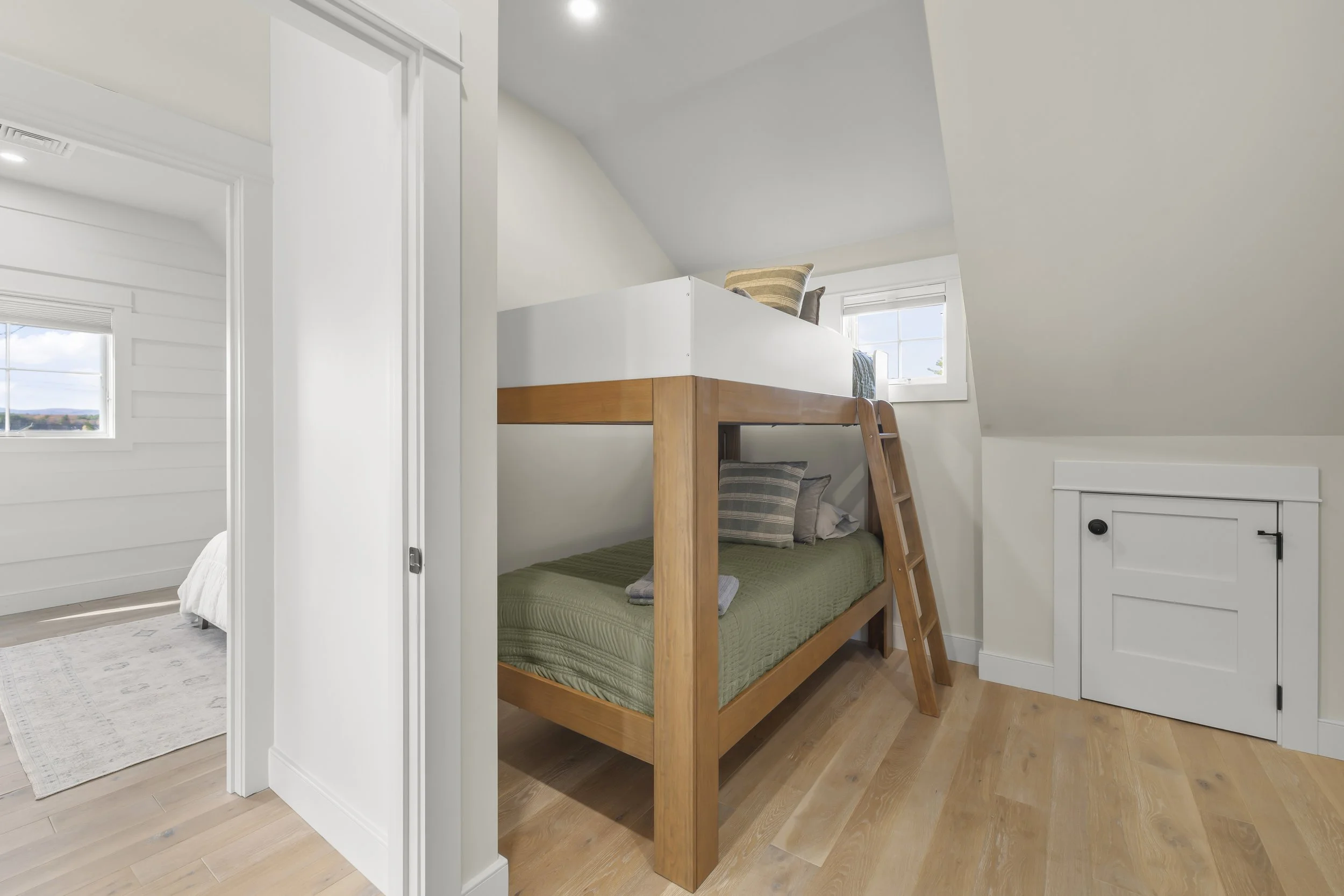 A wooden bunk bed with a ladder, situated in a small room with light hardwood floors and white walls, featuring a small storage closet and a window.