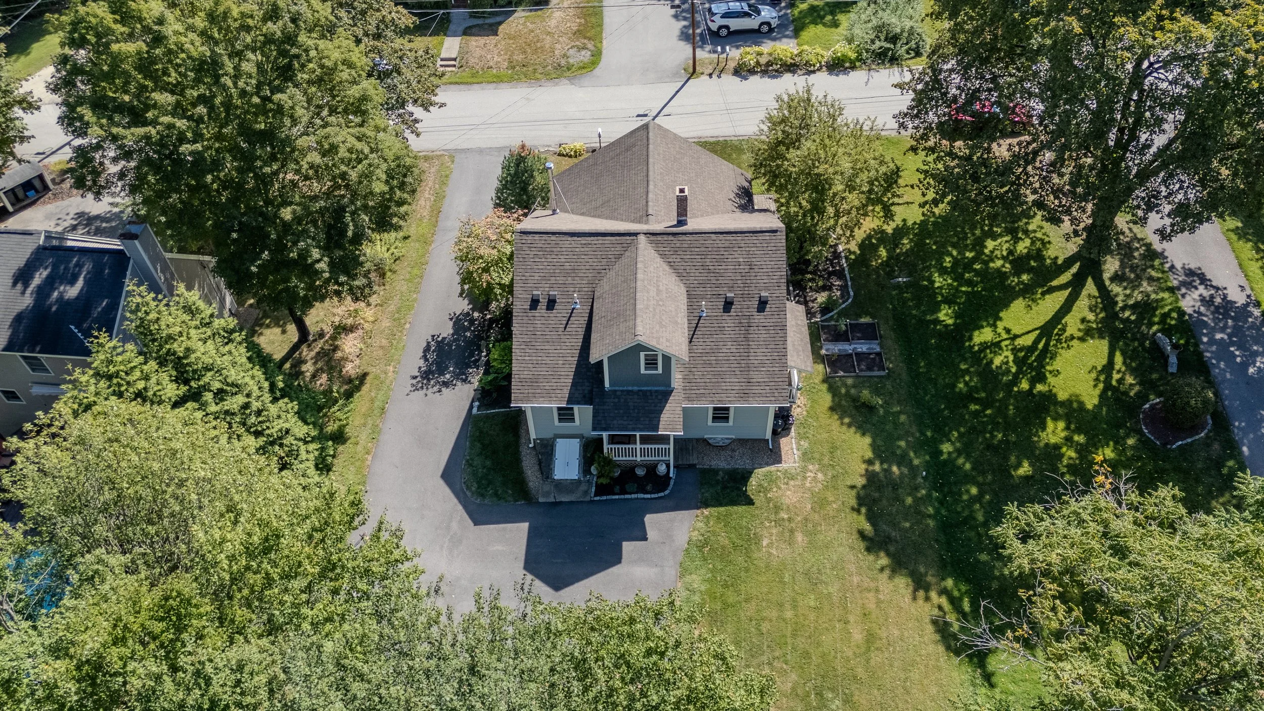 An aerial view of a house with a driveway, surrounded by green trees and lawn, with neighboring houses and a street visible.