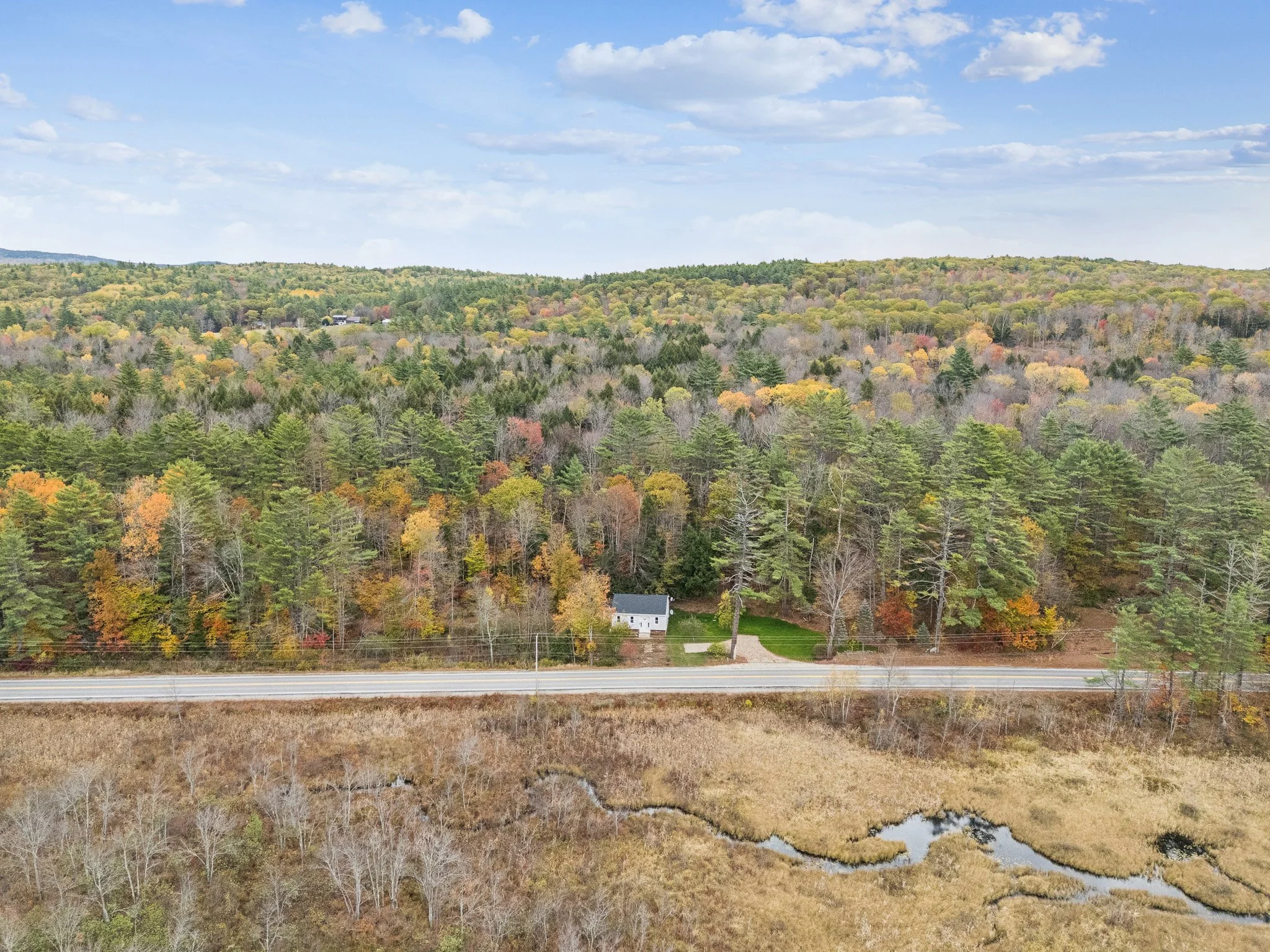 Aerial view of a rural landscape with a forest, a small white house, a road, and a wetland area in autumn.