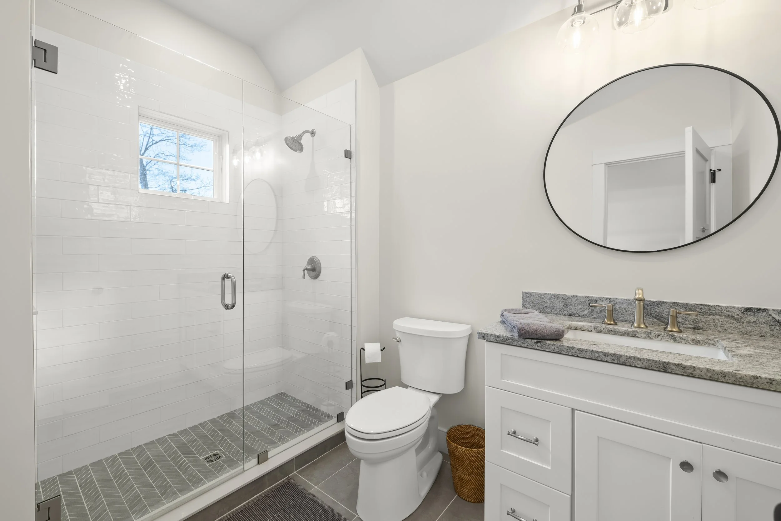 Modern white bathroom with a glass shower, a white toilet, and a vanity with a granite countertop and a round mirror.