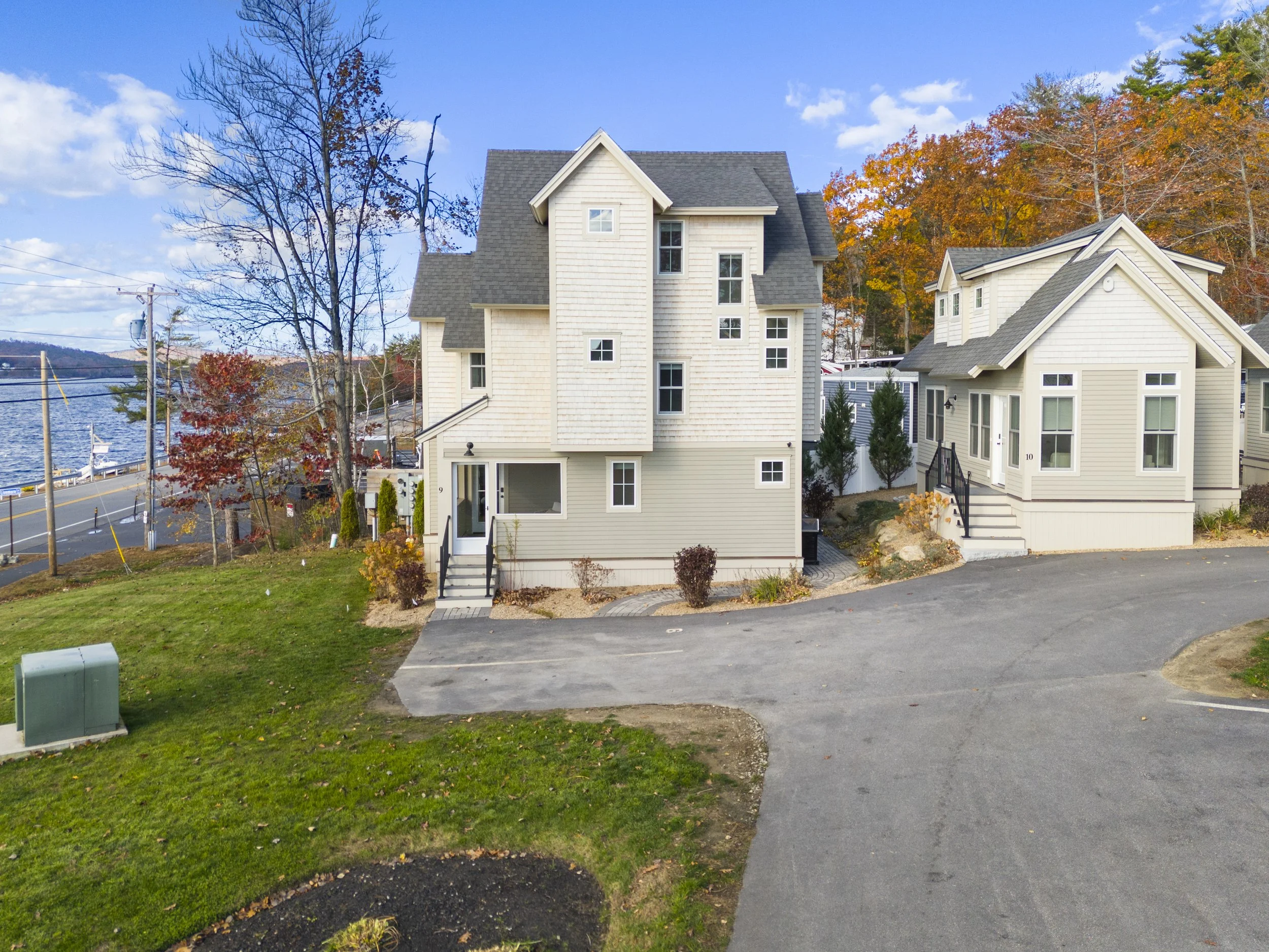 Two modern houses with white siding, set on a street near a body of water, with trees showing fall foliage, and a clear blue sky.