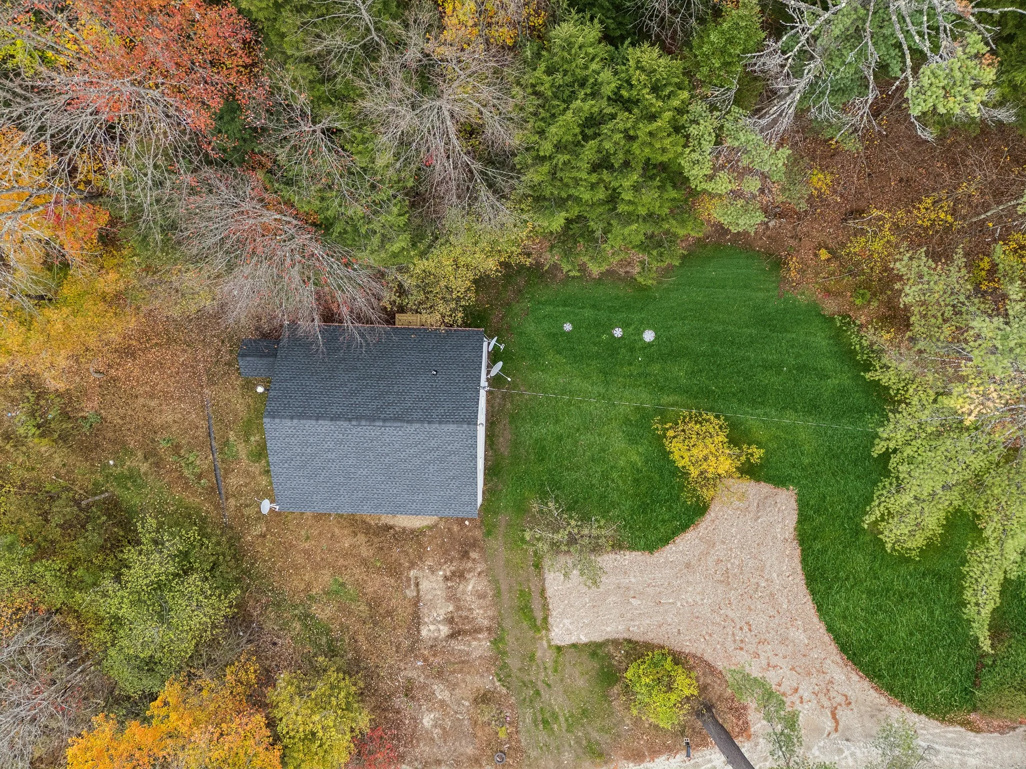 An aerial view of a house with a dark roof surrounded by trees with autumn foliage, green grass, and a dirt driveway.