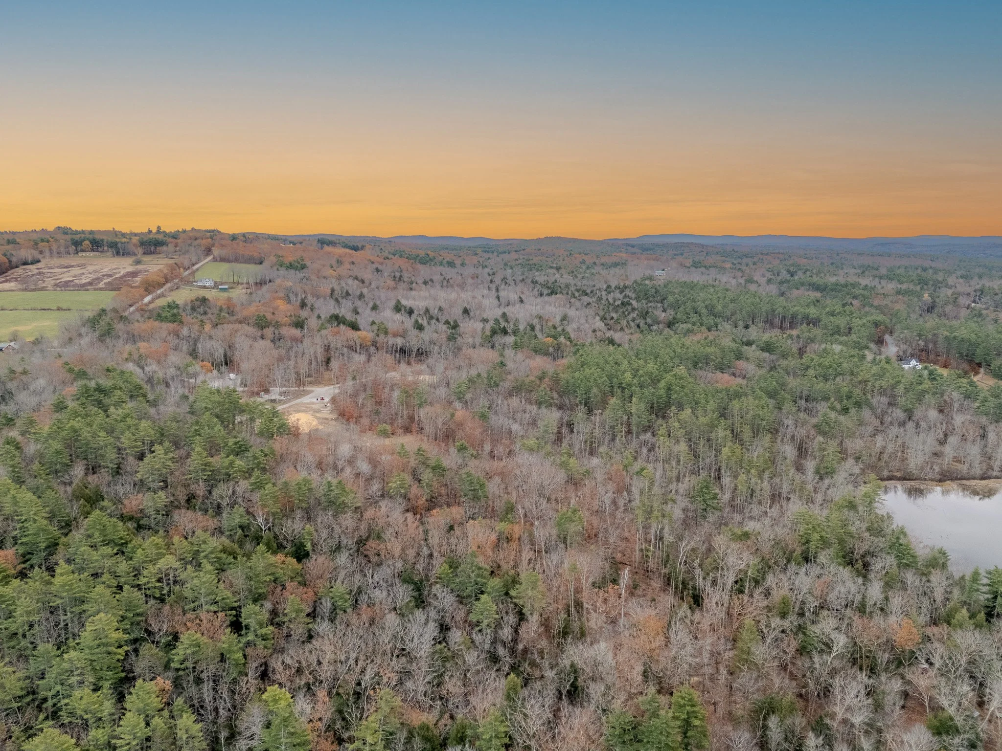 Aerial view of a forested landscape with a small pond to the right, fields, and scattered houses, under a sunset sky.