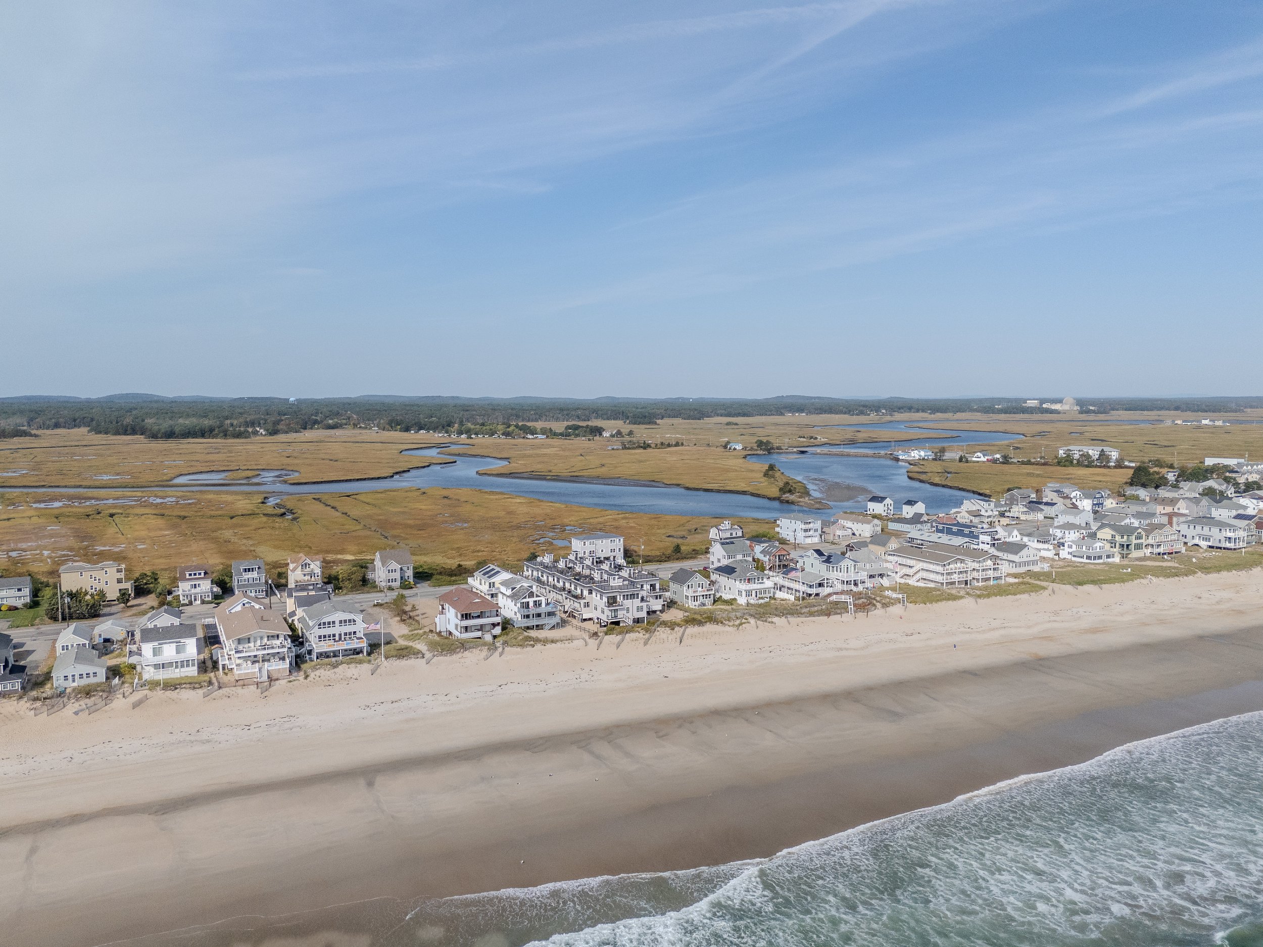 Aerial view of a coastal neighborhood with beach houses along the sandy shoreline, backed by marshland, waterways, and a blue sky.