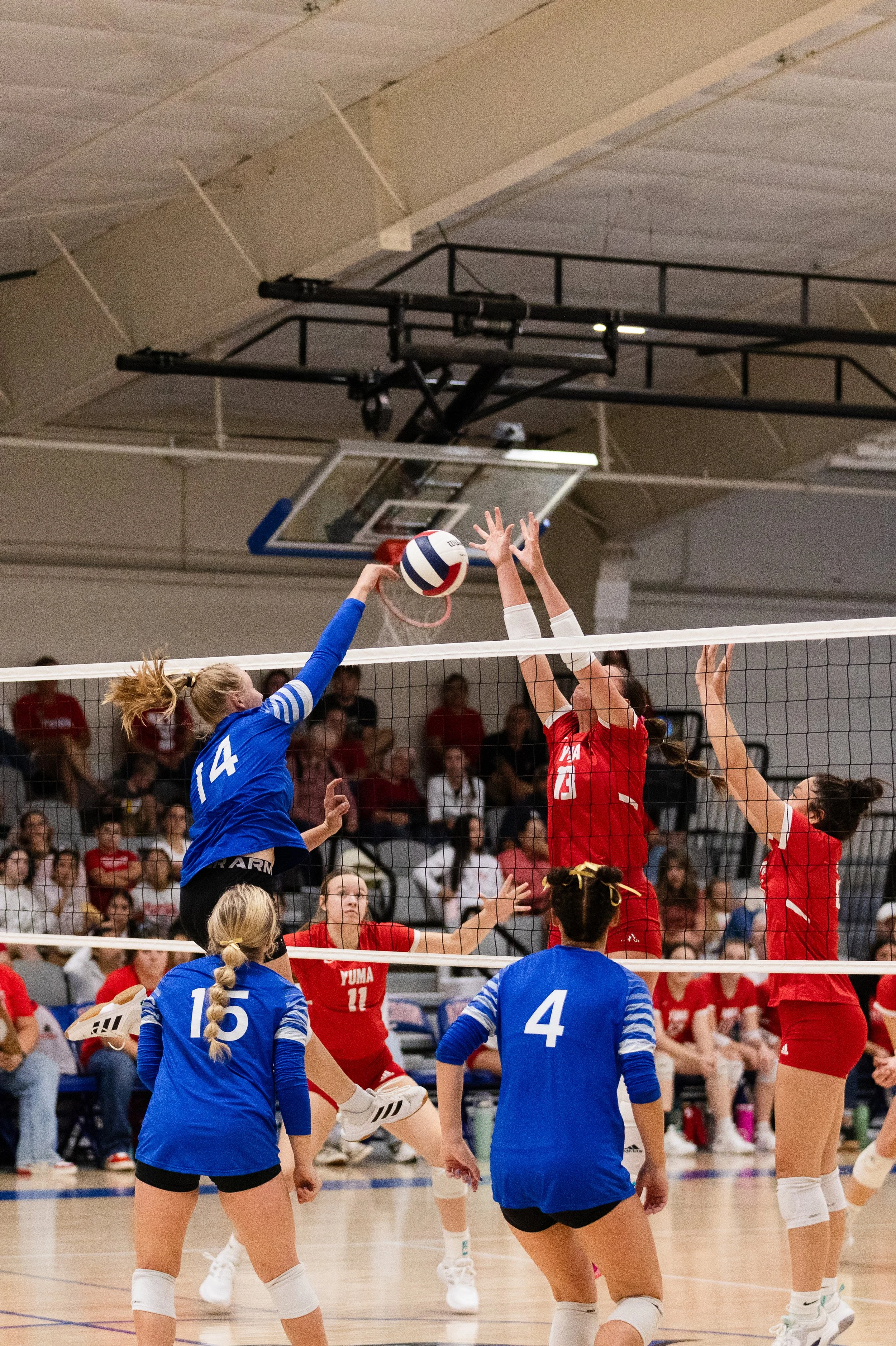 Women playing volleyball indoors, with some players jumping at the net and others preparing for the ball, in a gymnasium with a hardwood floor and some spectators in the background.