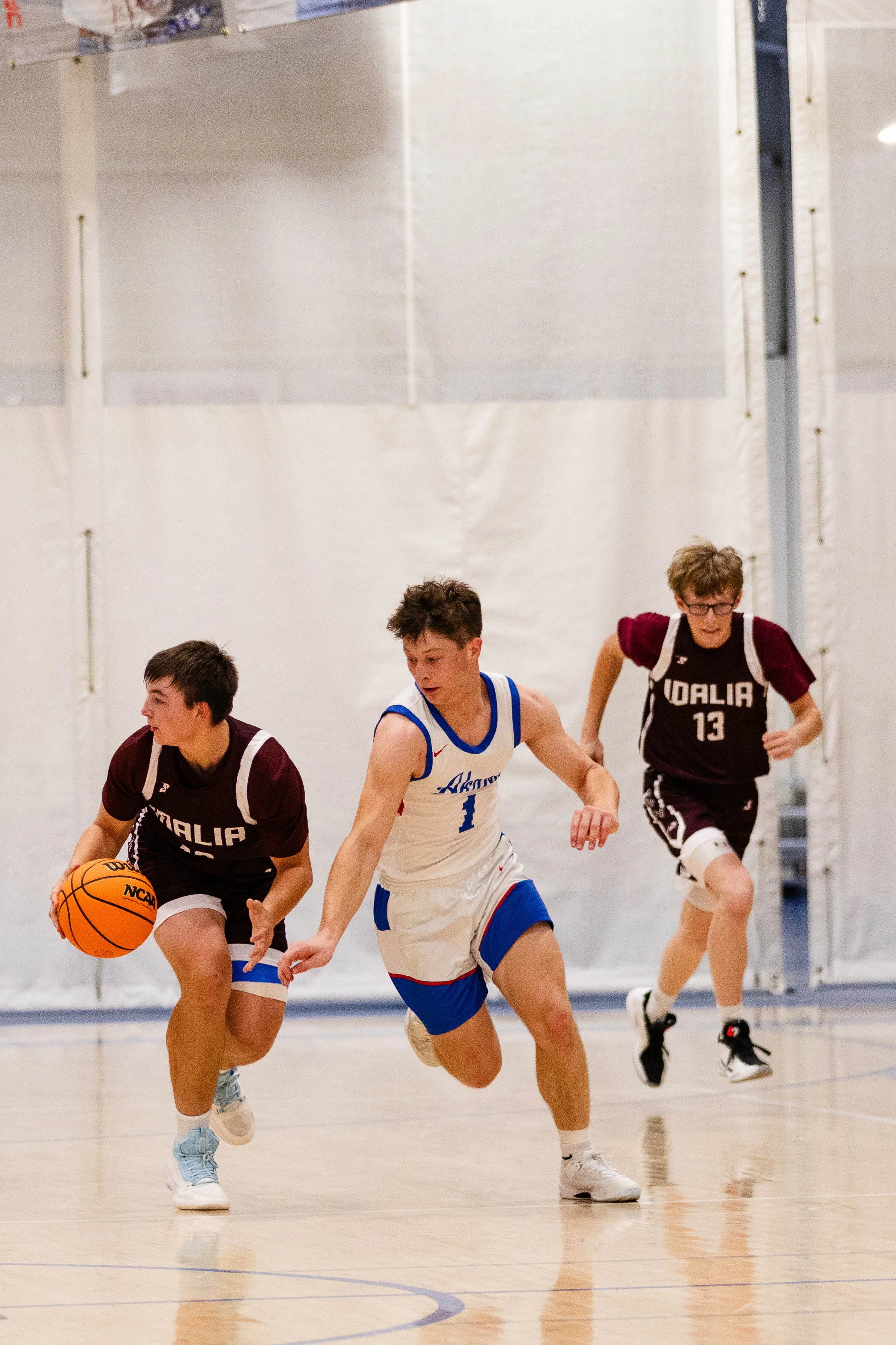 Three young male basketball players actively running on an indoor court during a game, with one dribbling the ball and the others pursuing him.