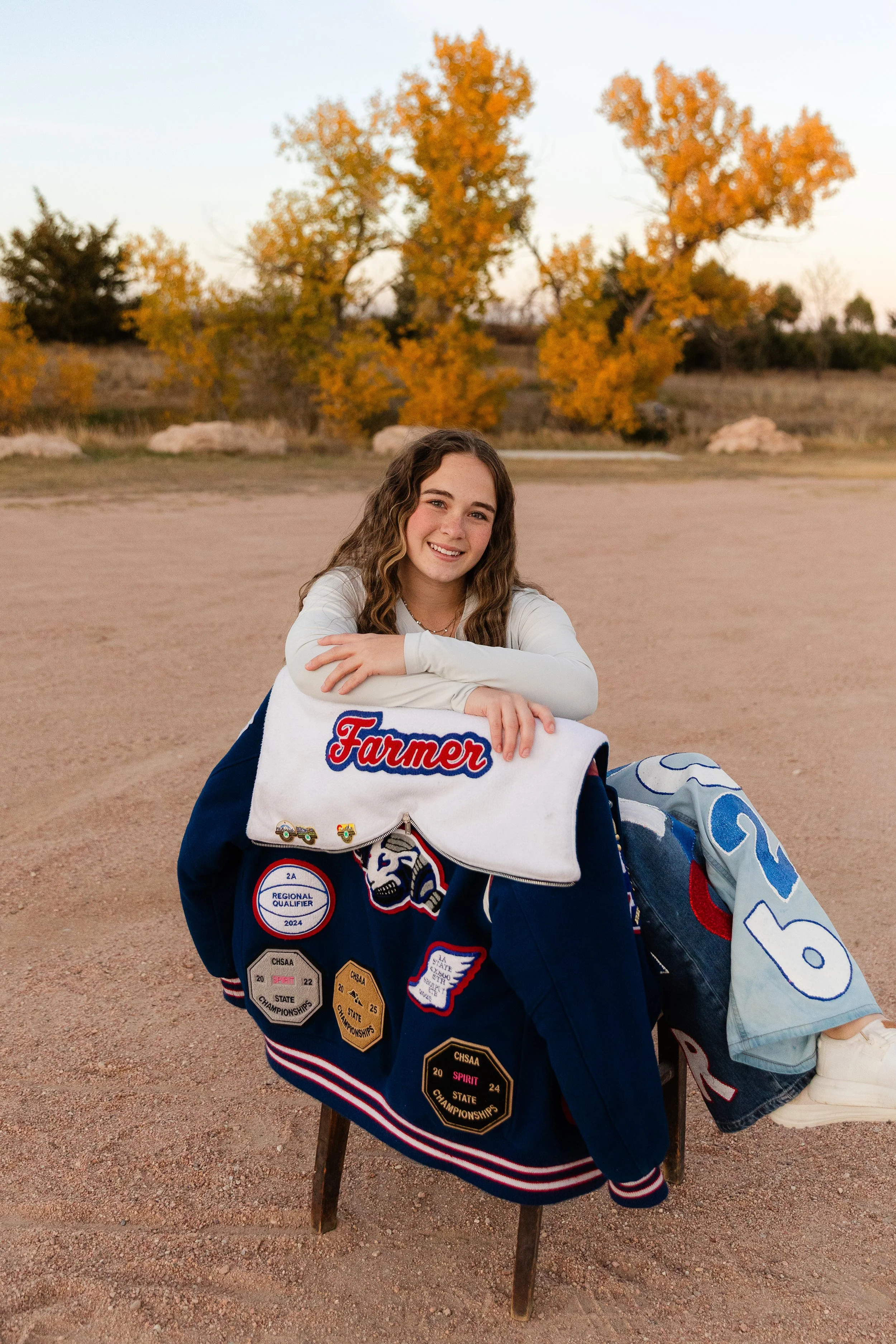 A young woman with long, wavy hair sits on a wooden stool on a dirt field during autumn. She is smiling and has her arms resting on a clothing rack covered with various sports patches and badges, including one that says 'Farmer.' She is wearing a white long-sleeve shirt and gray shorts, with a hockey stick on the ground next to her. The background features trees with vibrant orange and yellow leaves.