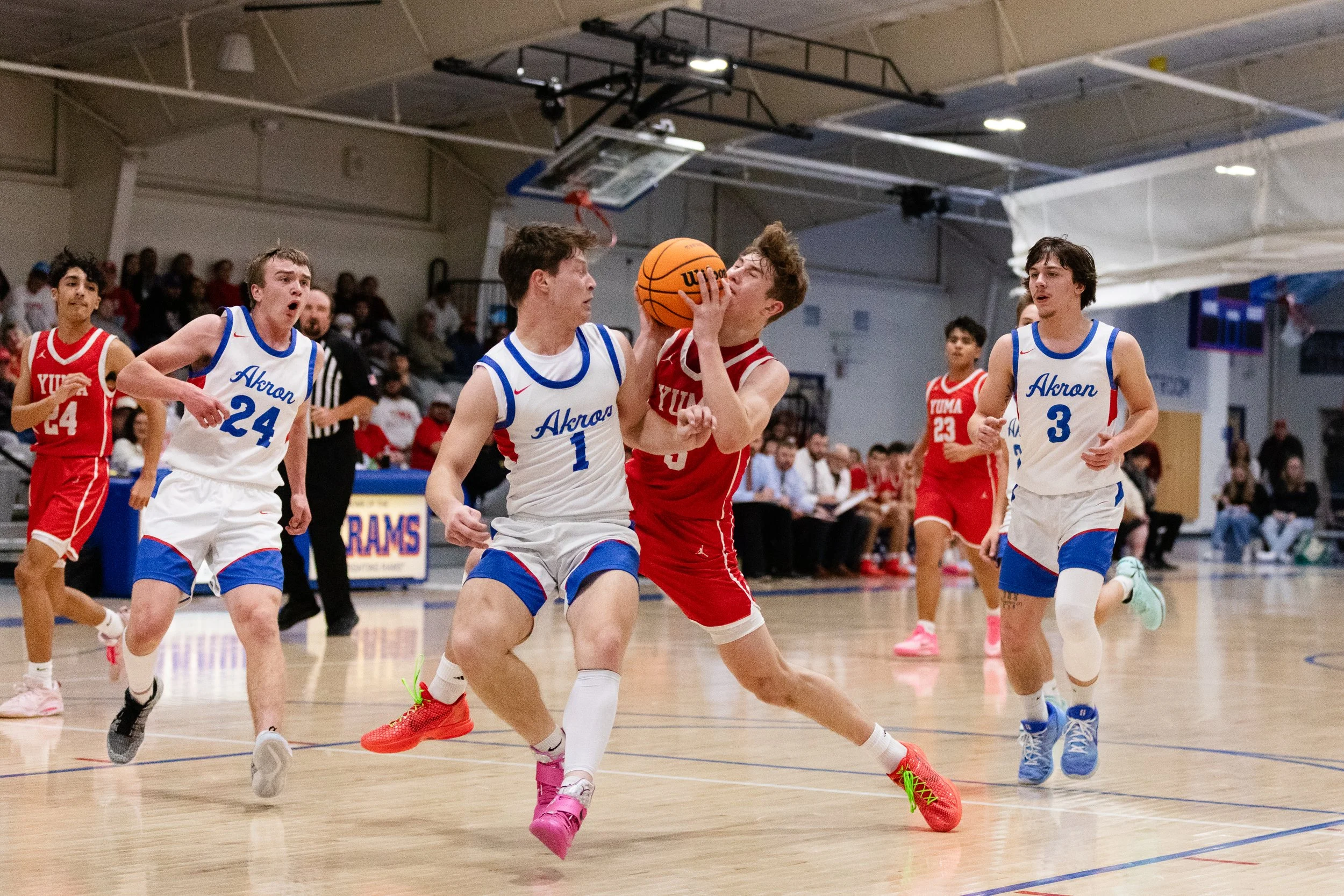 Young basketball players in red and white jerseys playing a game indoors. One player is attempting a jump shot while another is defending close by. Spectators are seated in the background.
