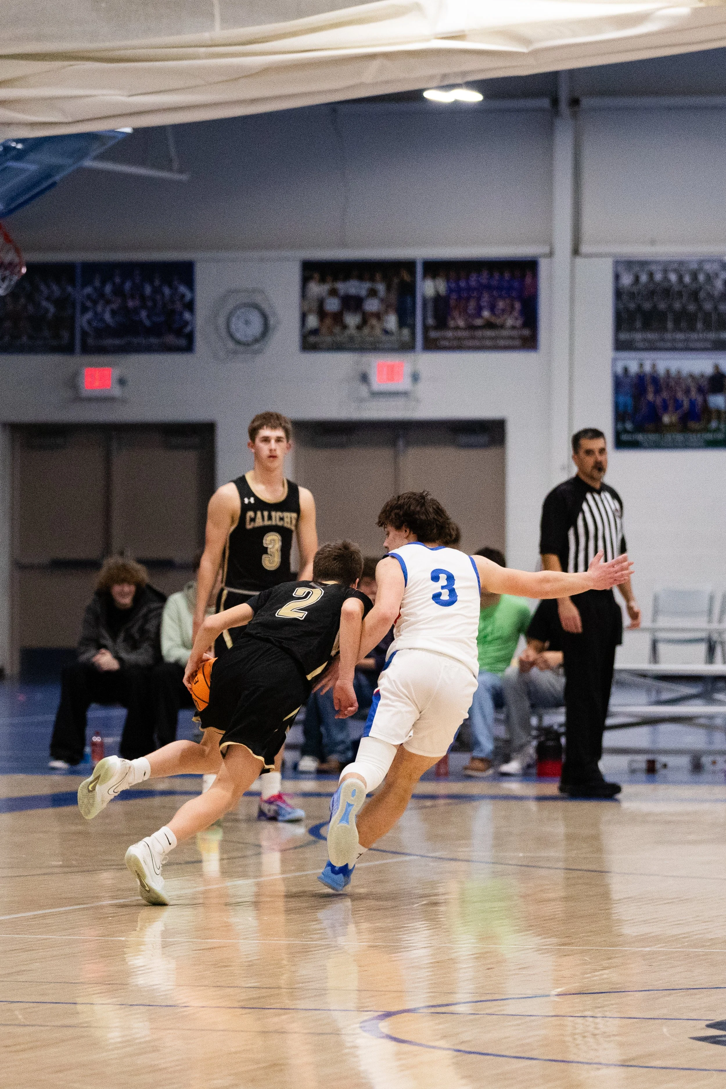 Two basketball players in jerseys, one in black and the other in white with blue accents, competing for the ball on an indoor basketball court. A referee and spectators are visible in the background.