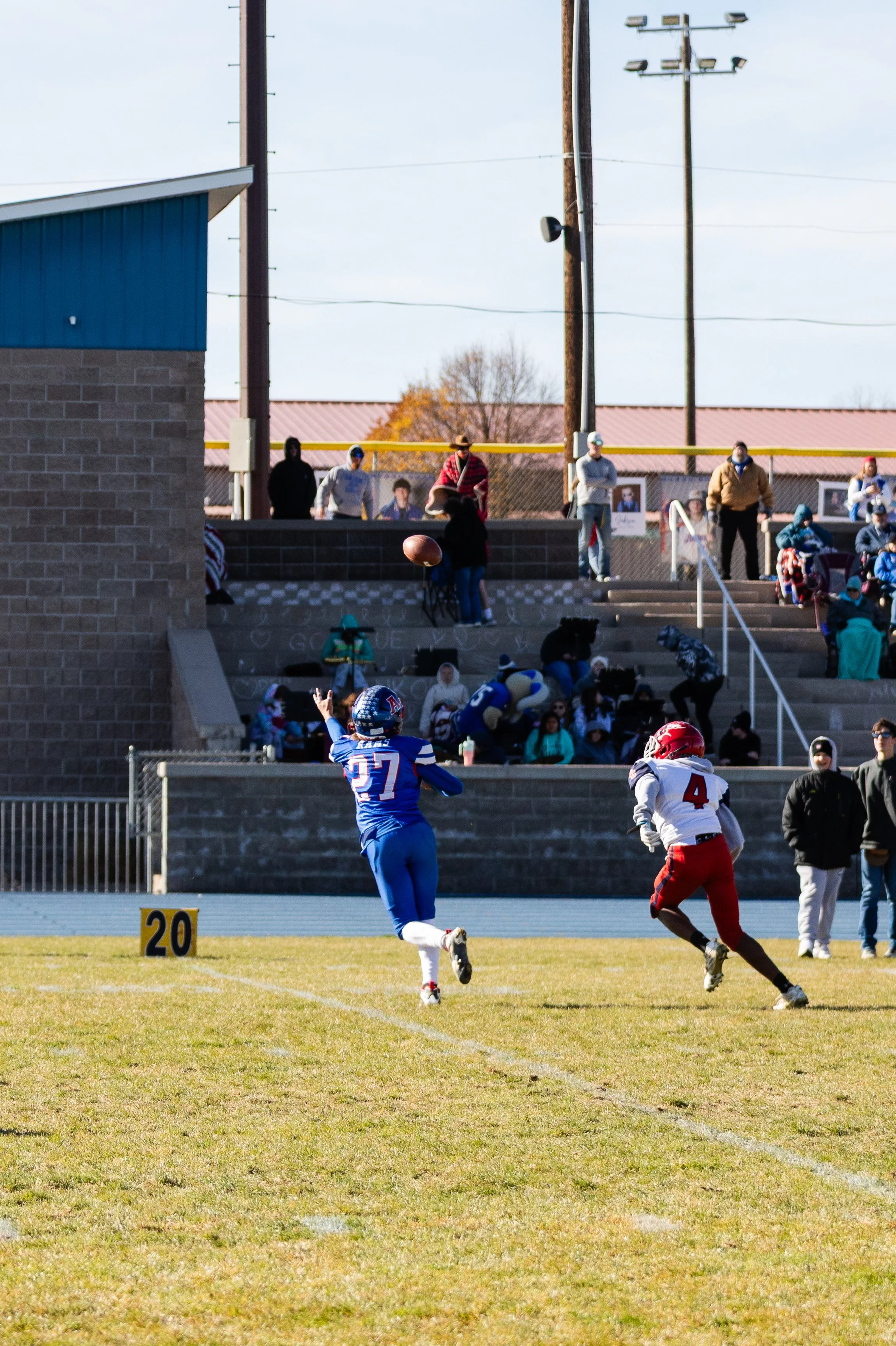 A football game in progress with two players on the field, one in a blue uniform and another in a white and red uniform, near the 20-yard line. Spectators are watching from the stands with some taking photos, and a person is holding a football. The b