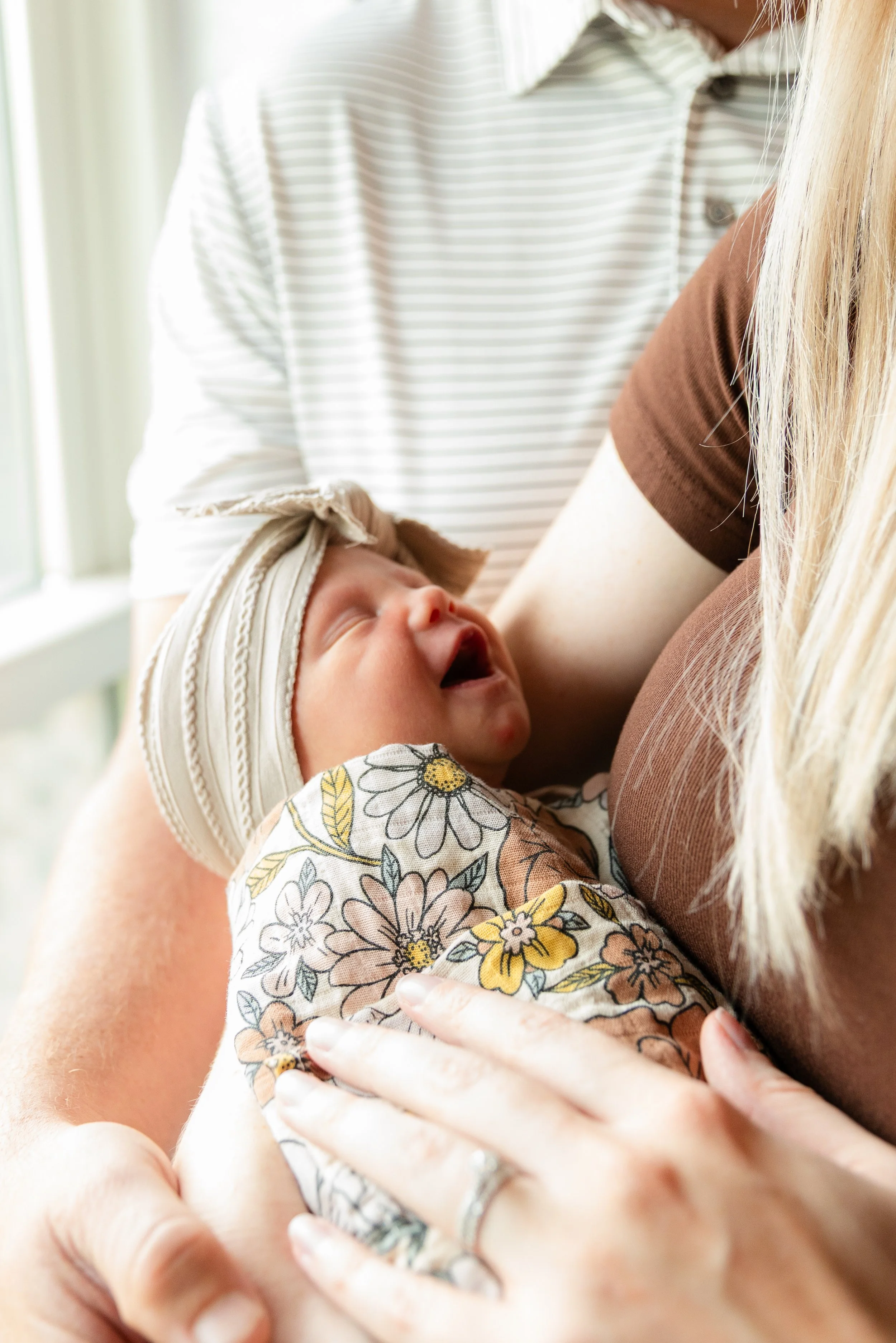 A person is holding a yawning sleeping baby against their chest, with another person nearby.