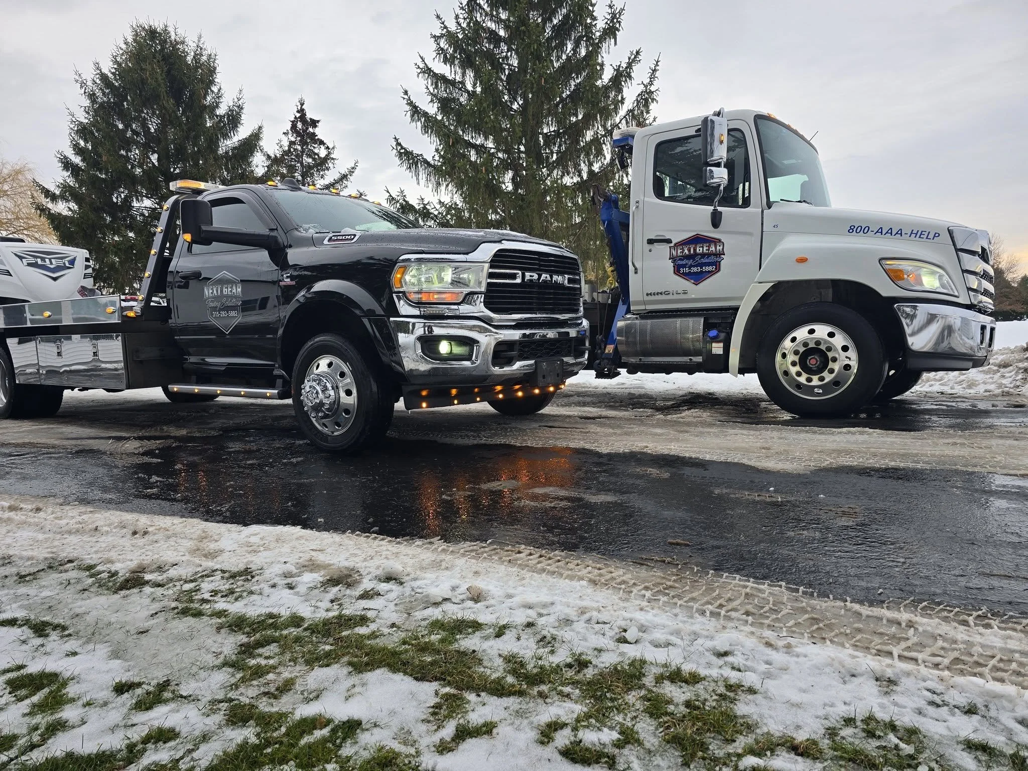 A black Dodge truck connected to a white Kenworth tow truck on a snowy and muddy ground with evergreen trees in the background.