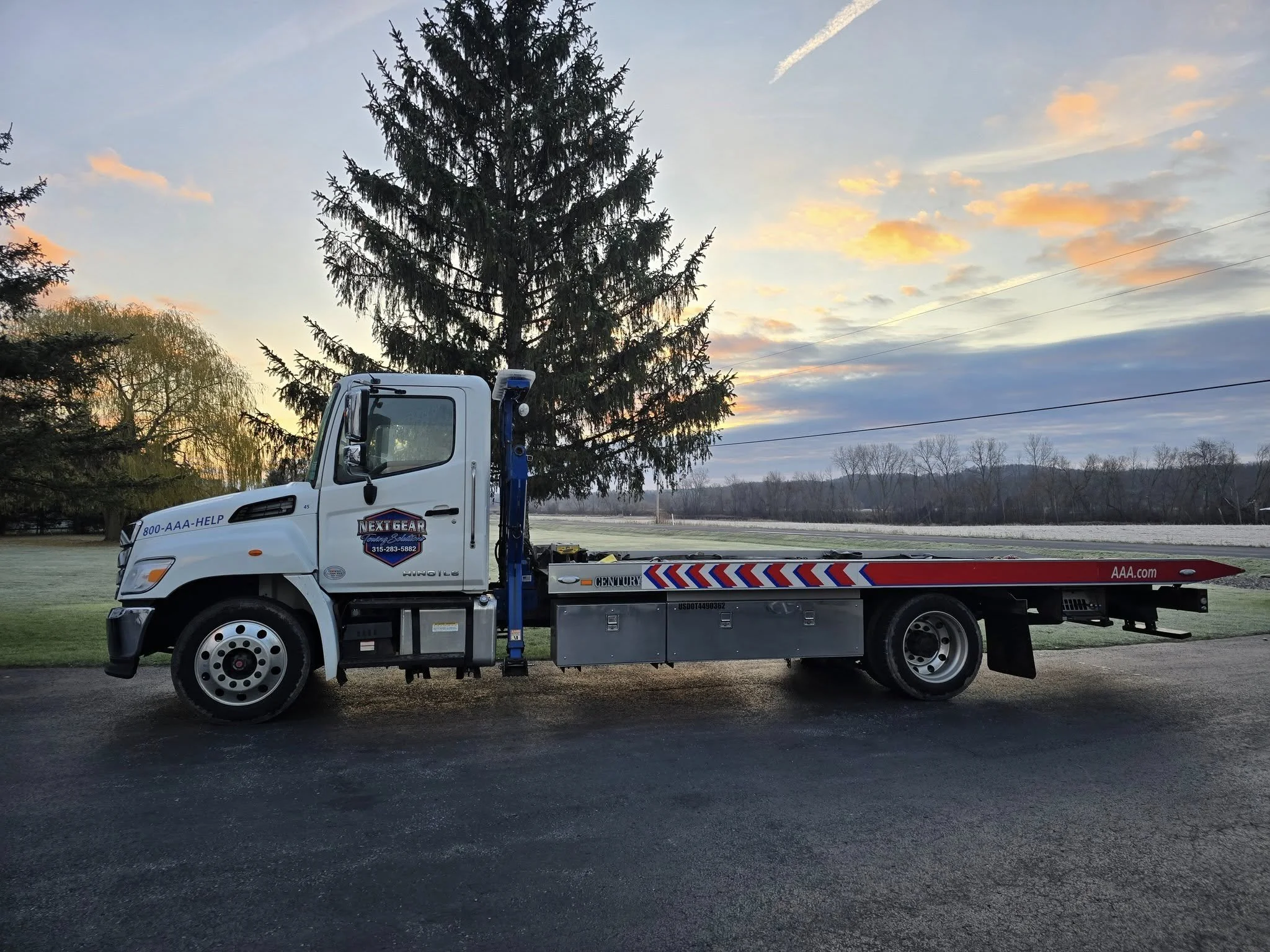 A flatbed tow truck parked on a paved surface with trees and a sunset sky in the background.