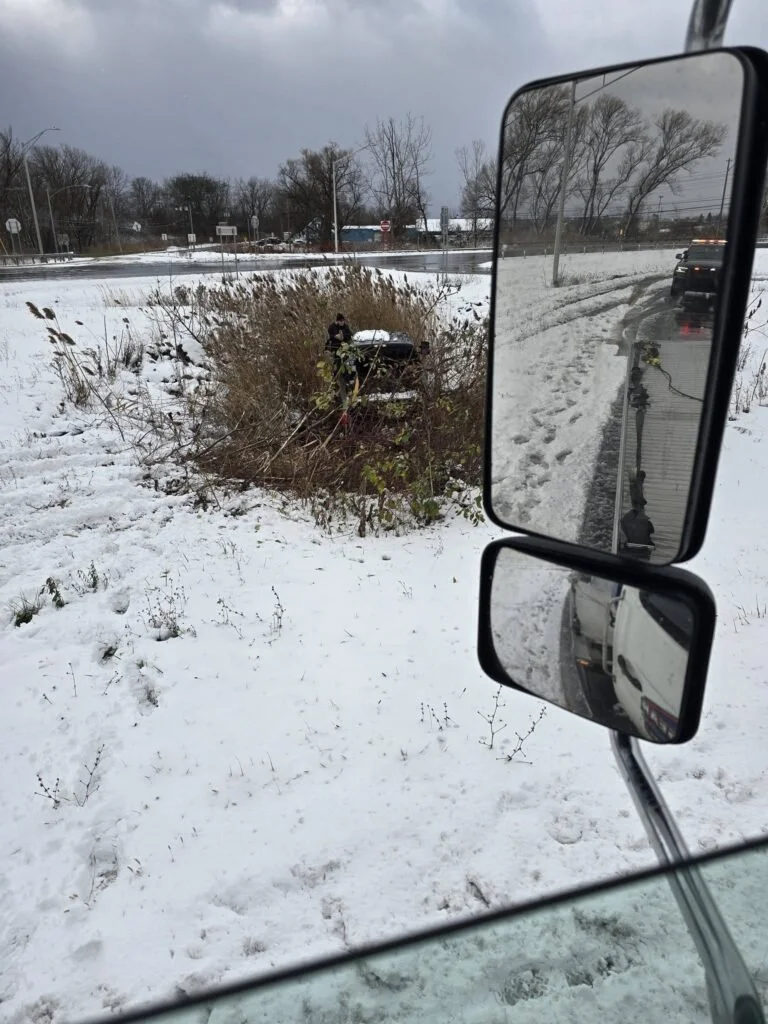 Vehicle accident in a snowy area with a car stuck in bushes, seen from the side mirror of a truck.