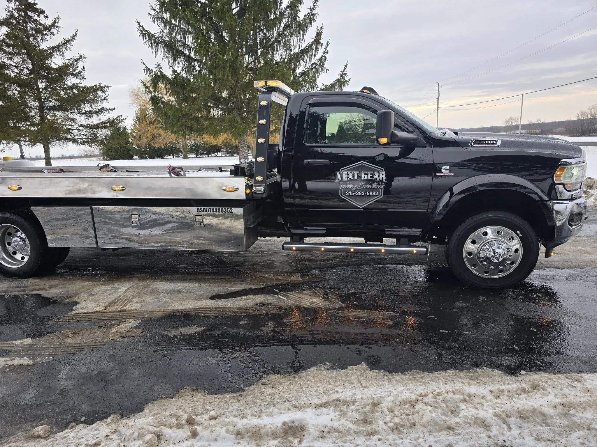 Black tow truck with Next Gear logo parked on wet asphalt in winter with snow and trees in the background.